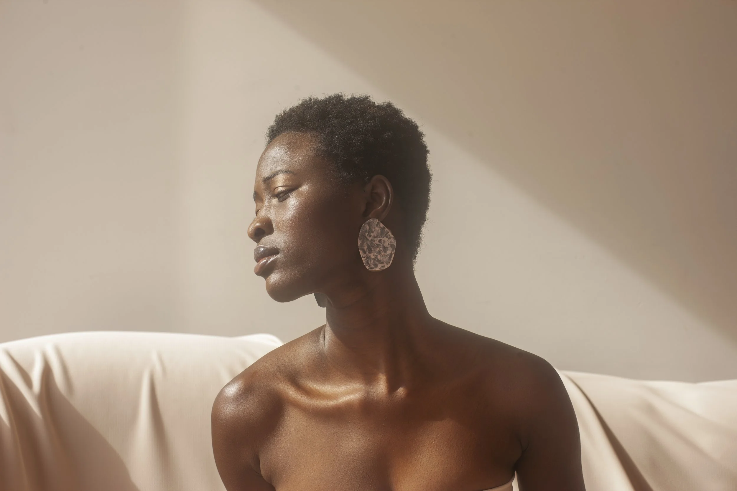 A Black woman with short curly hair, wearing large patterned earrings, is sitting against a neutral background with soft lighting, looking downward with a serene expression.