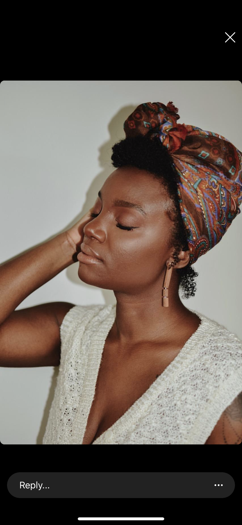 A woman with eyes closed, wearing a colorful patterned headwrap and a knitted sleeveless top, standing against a plain background wearing earrings made from old violin bows.