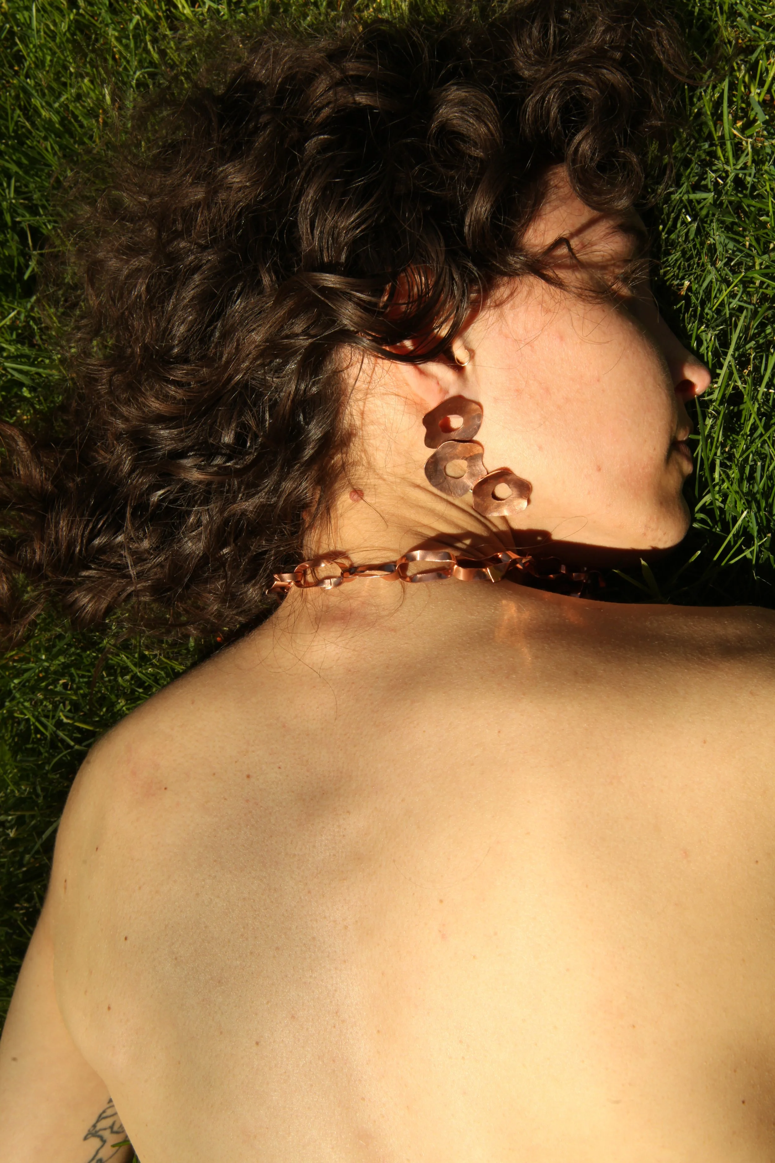 A woman with curly brown hair lying on green grass, wearing copper earrings and a necklace, with her eyes closed and face turned to the side.