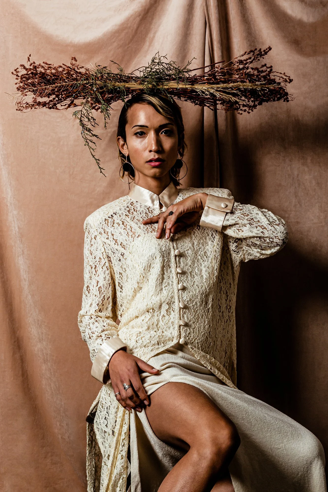 A woman wearing a cream lace blouse with pearl buttons, sitting against a beige backdrop. She has a dried floral arrangement balanced on her head, and her pose includes one hand near her face and the other on her lap wearing large statement earrings.