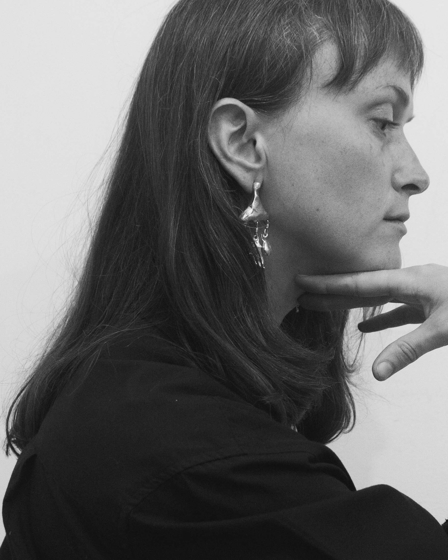 A woman with long dark hair wearing earrings, resting her chin on her hand, in profile, in black and white.