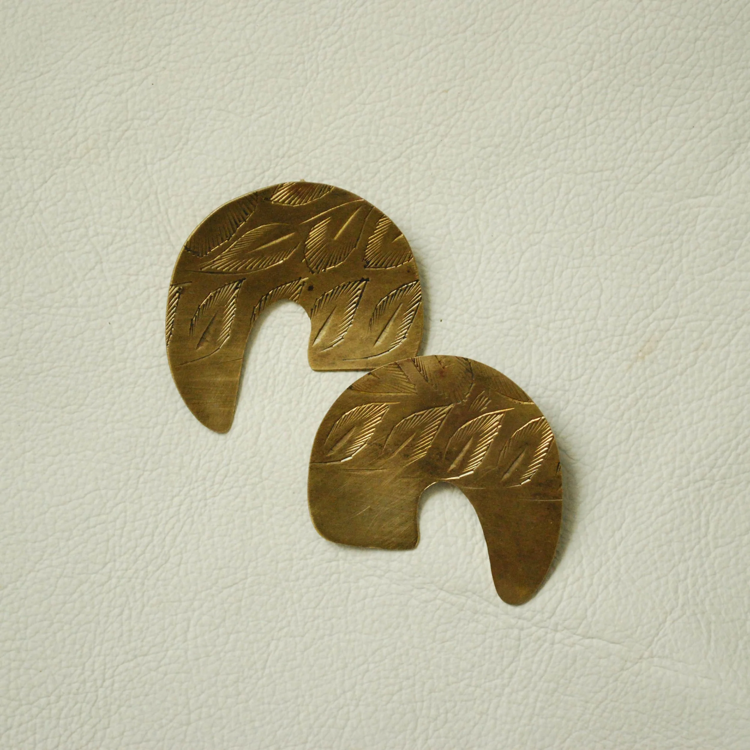 Two brass earrings with engraved leaf patterns on a white textured surface.