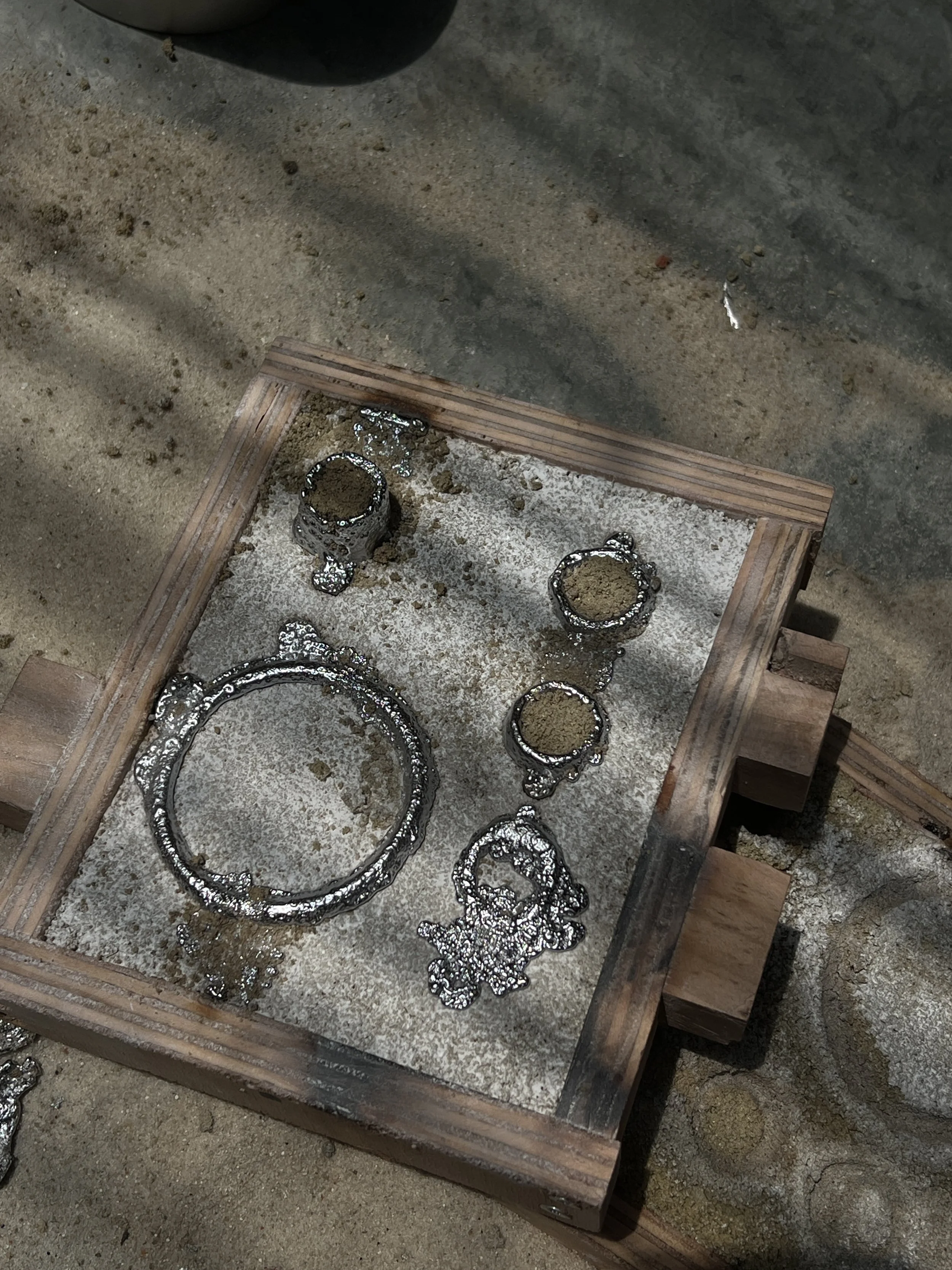 Metal rings and parts in a wooden mold, with sand and molten metal, on a concrete surface.
