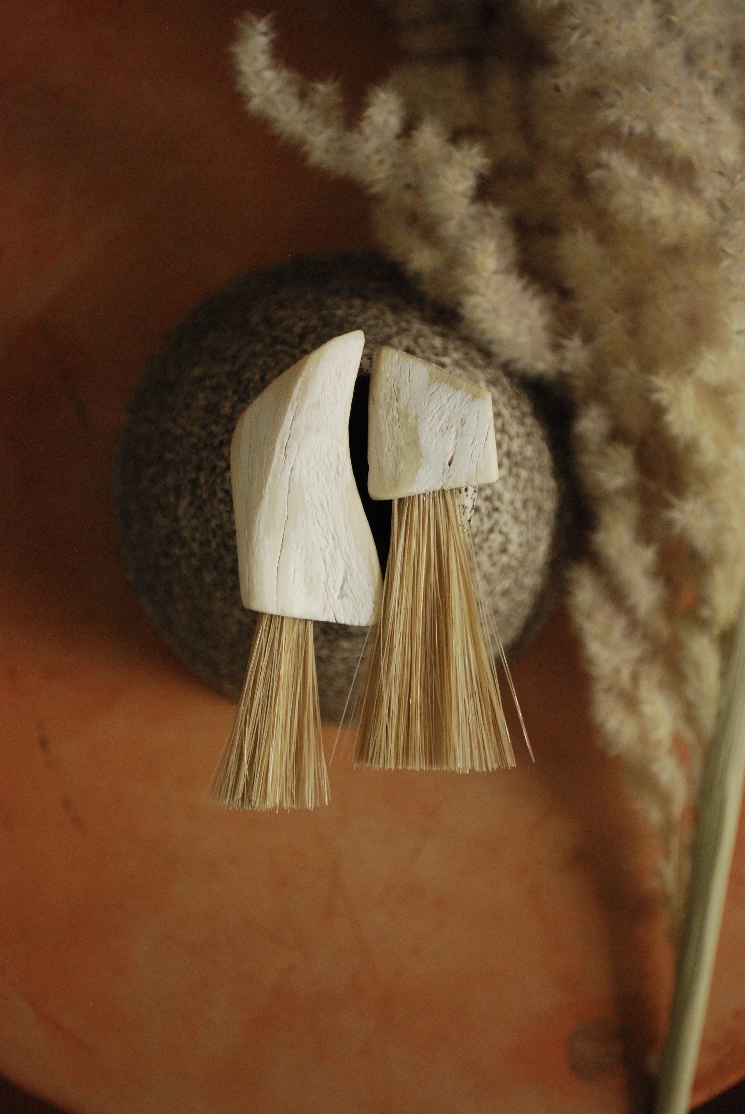 Pair of modern bone earrings with elk fur tassels, placed on a textured brown surface with soft beige and brown fabrics nearby.