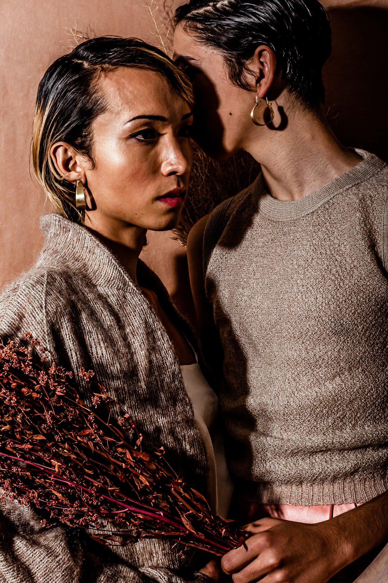 Two women with short dark hair and hoop earrings pose close together indoors, one holding a bouquet of dried flowers, both dressed in neutral tones.