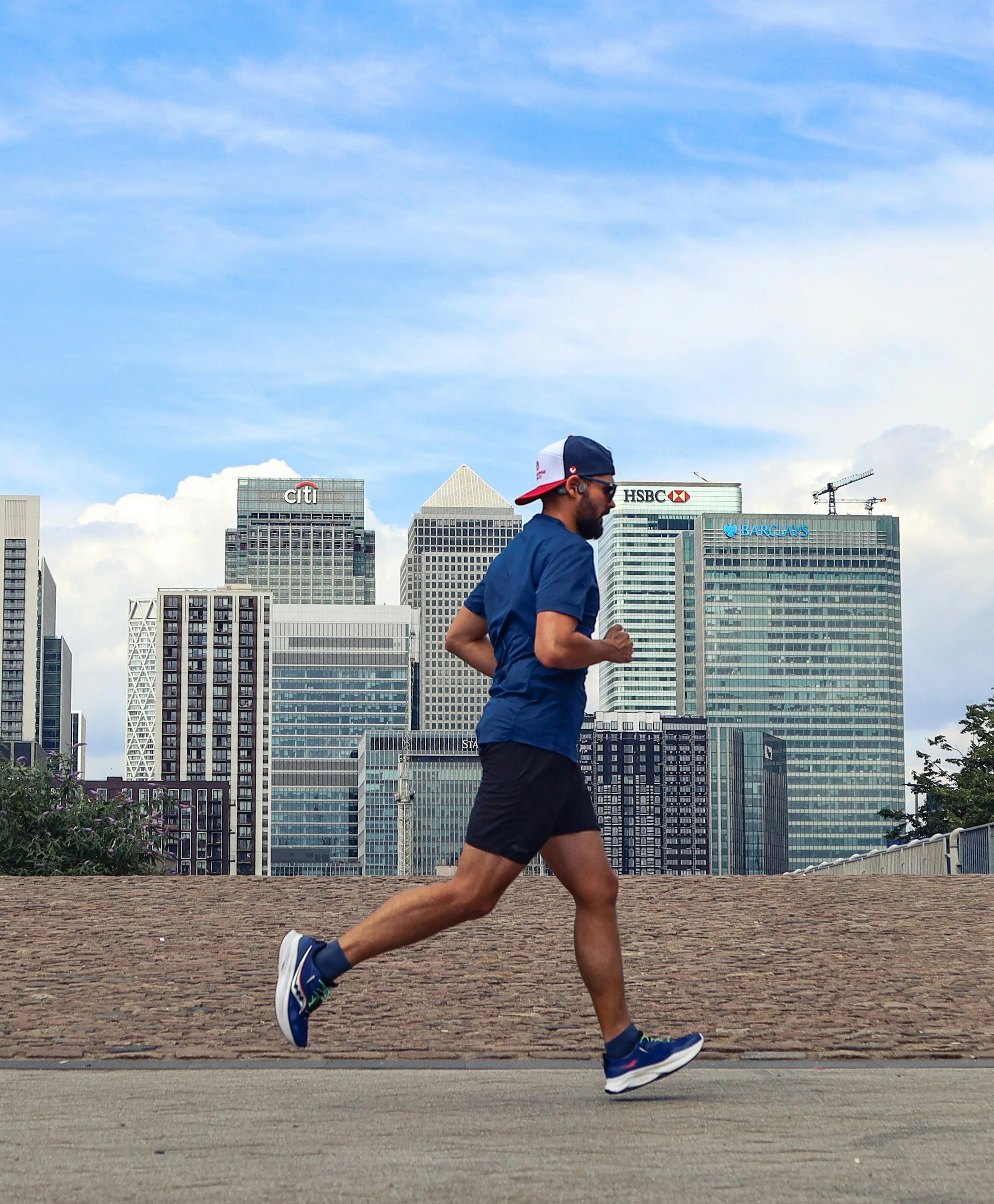 A man running in athletic wear on a paved path with a city skyline in the background, including skyscrapers with visible logos of Citi, HSBC, and Barclays.