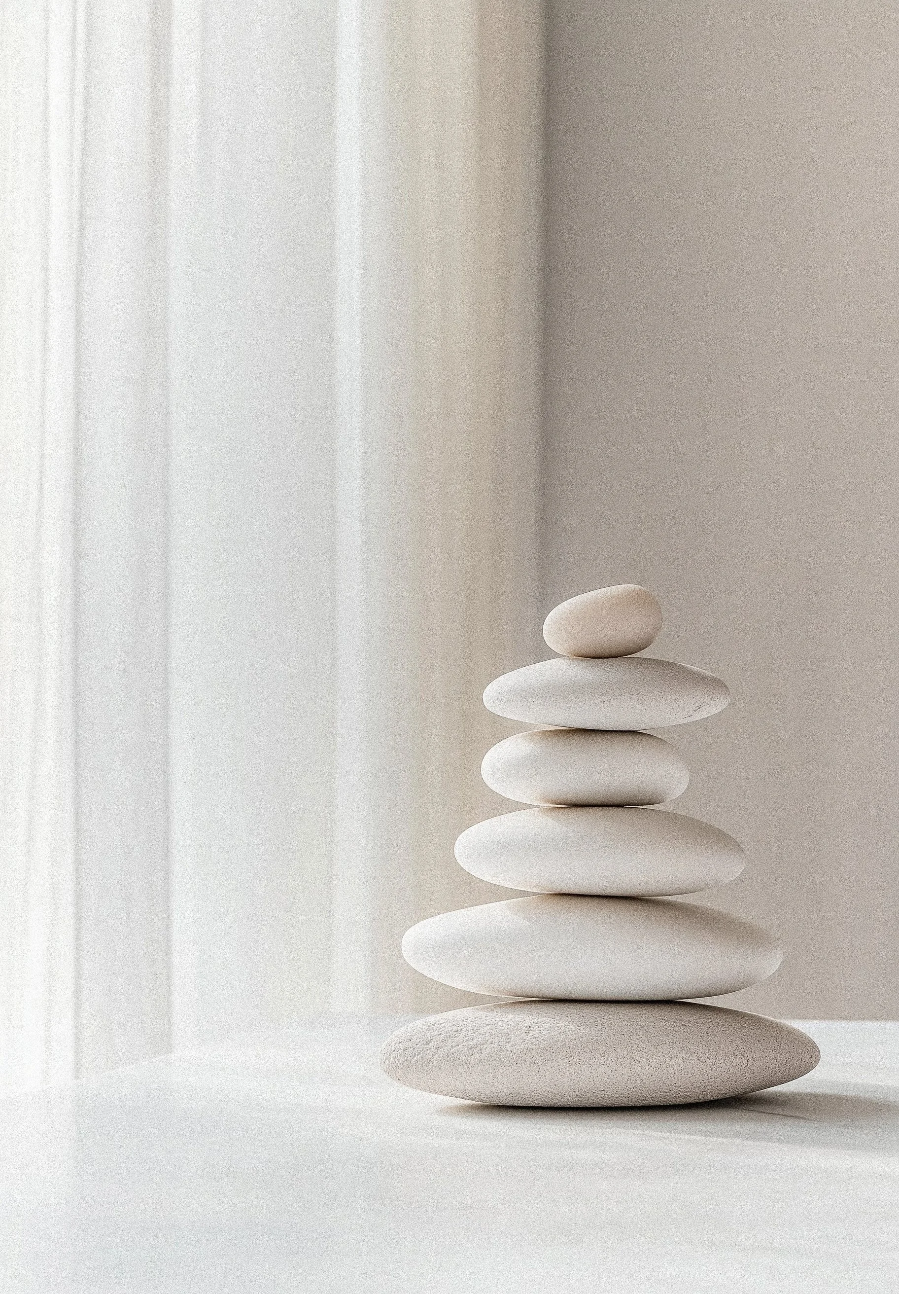 A stack of smooth white rocks arranged from largest at the bottom to smallest at the top on a white surface near a window with light filtering through.