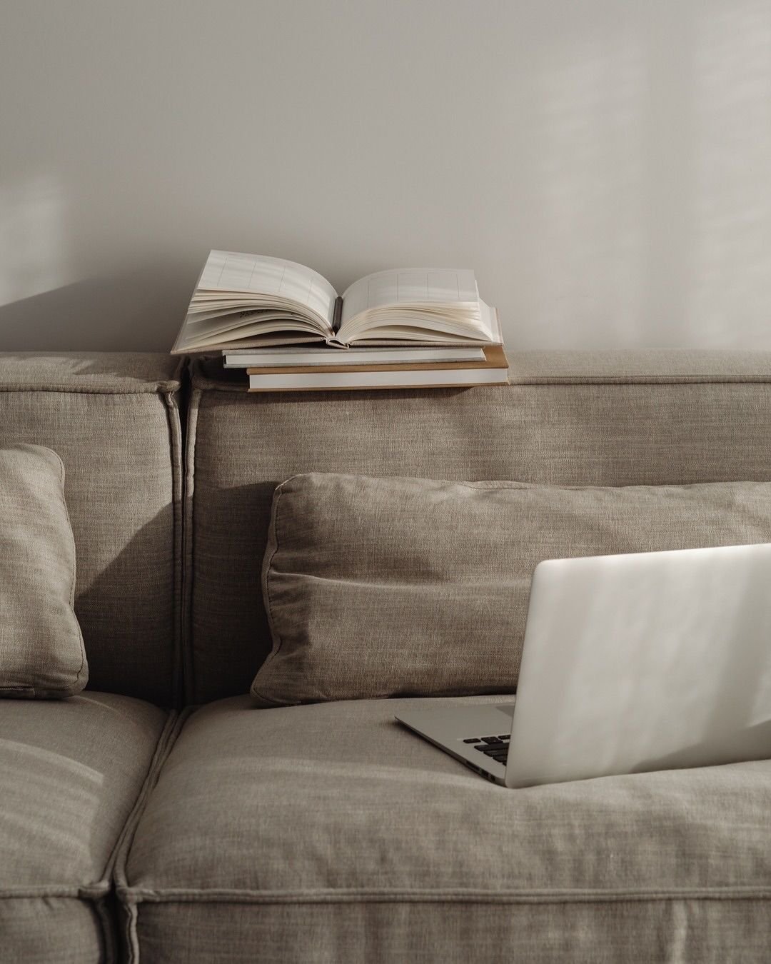 A beige fabric sofa with a laptop on the right side and an open book on top of two closed books on the backrest, against a plain white wall.