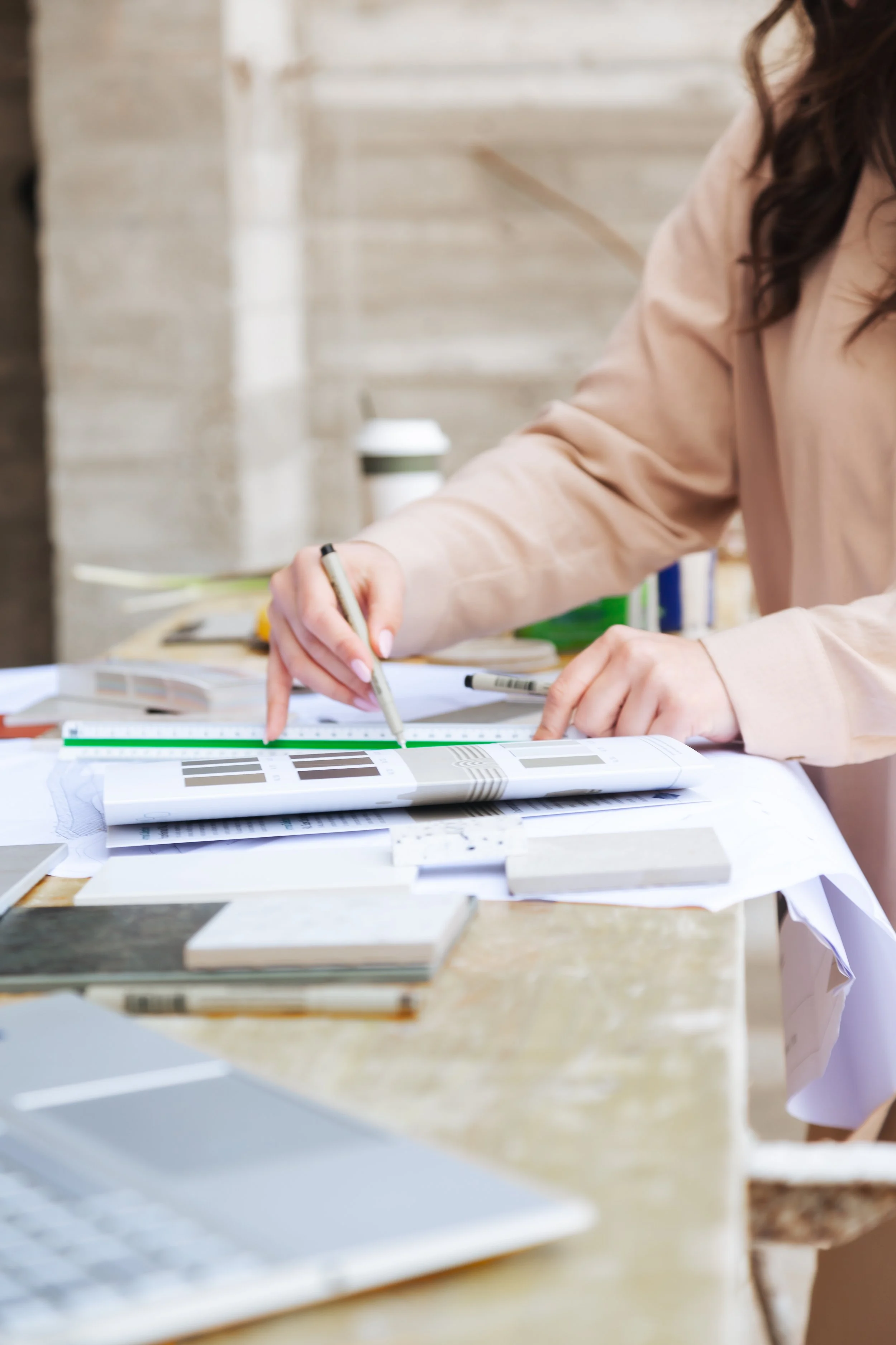 A person in a beige coat working on interior design plans with color swatches, a ruler, pens, and a laptop on a wooden table.