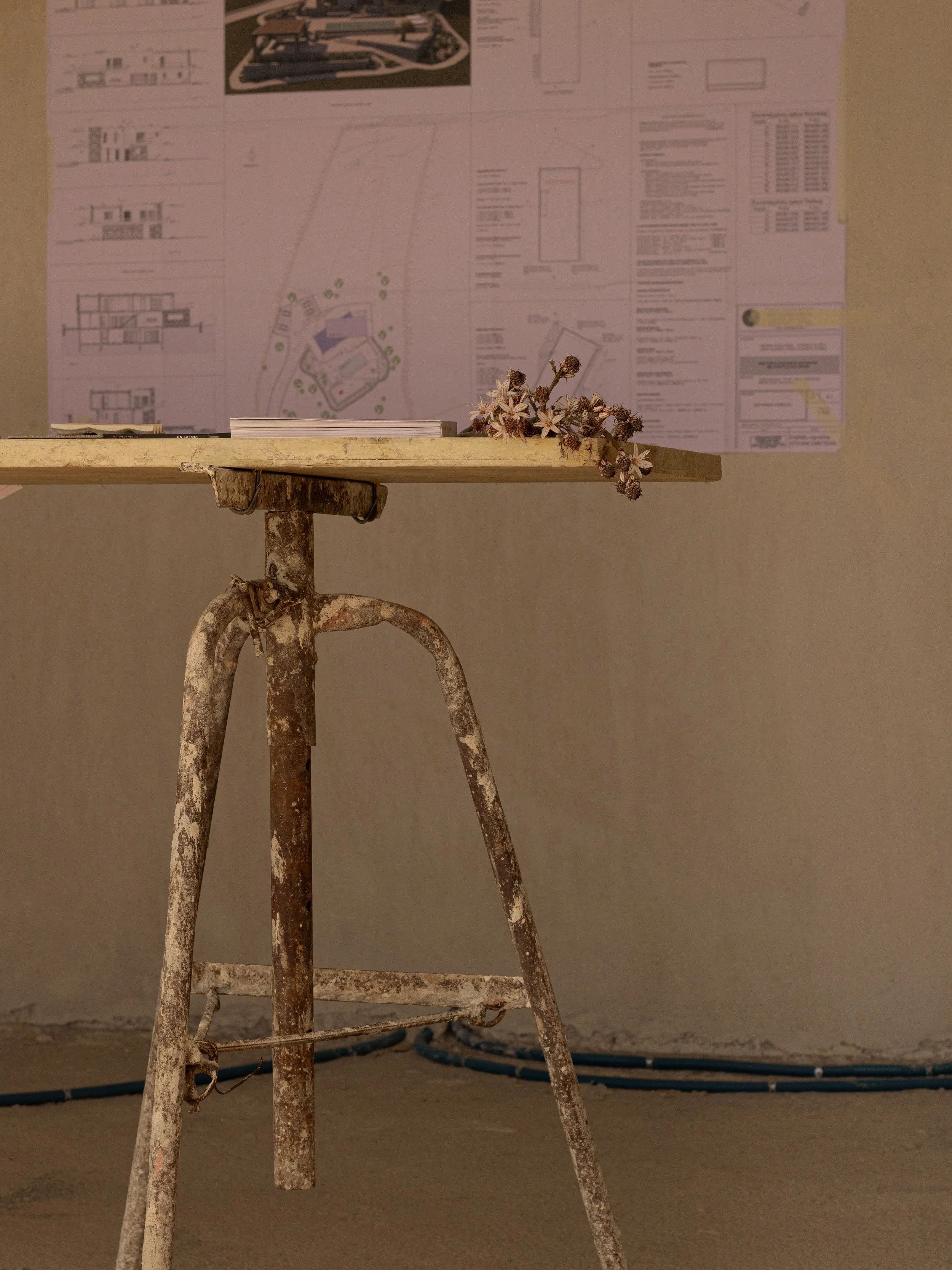 Old, rusty metal stool with a wooden top, situated in front of a wall with architectural plans taped to it. There are some dried flowers on the stool's edge.