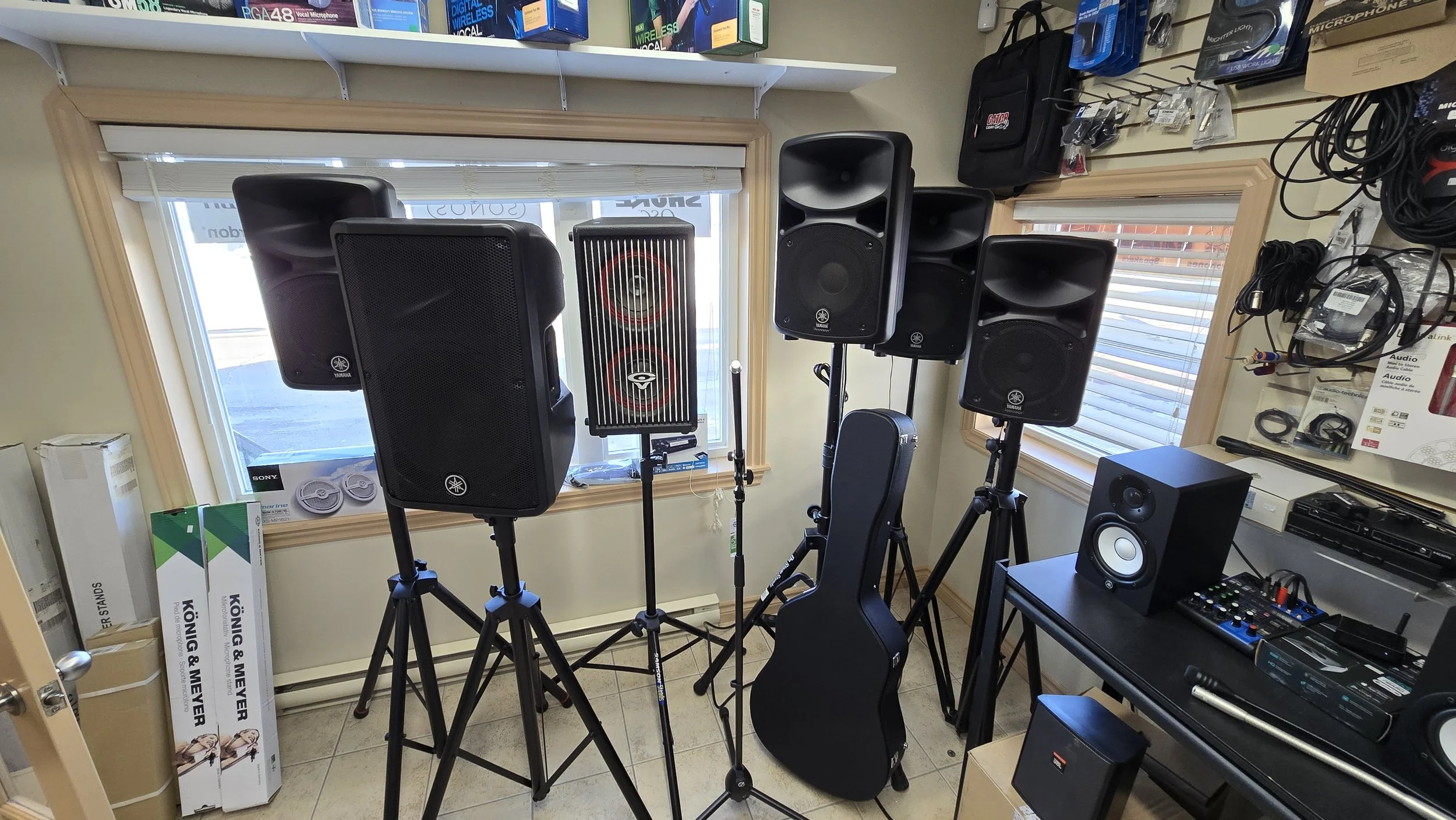 A music equipment store display with several Yamaha speakers on stands, a guitar in a black case, a mixing console, and various audio accessories on wall and shelves.