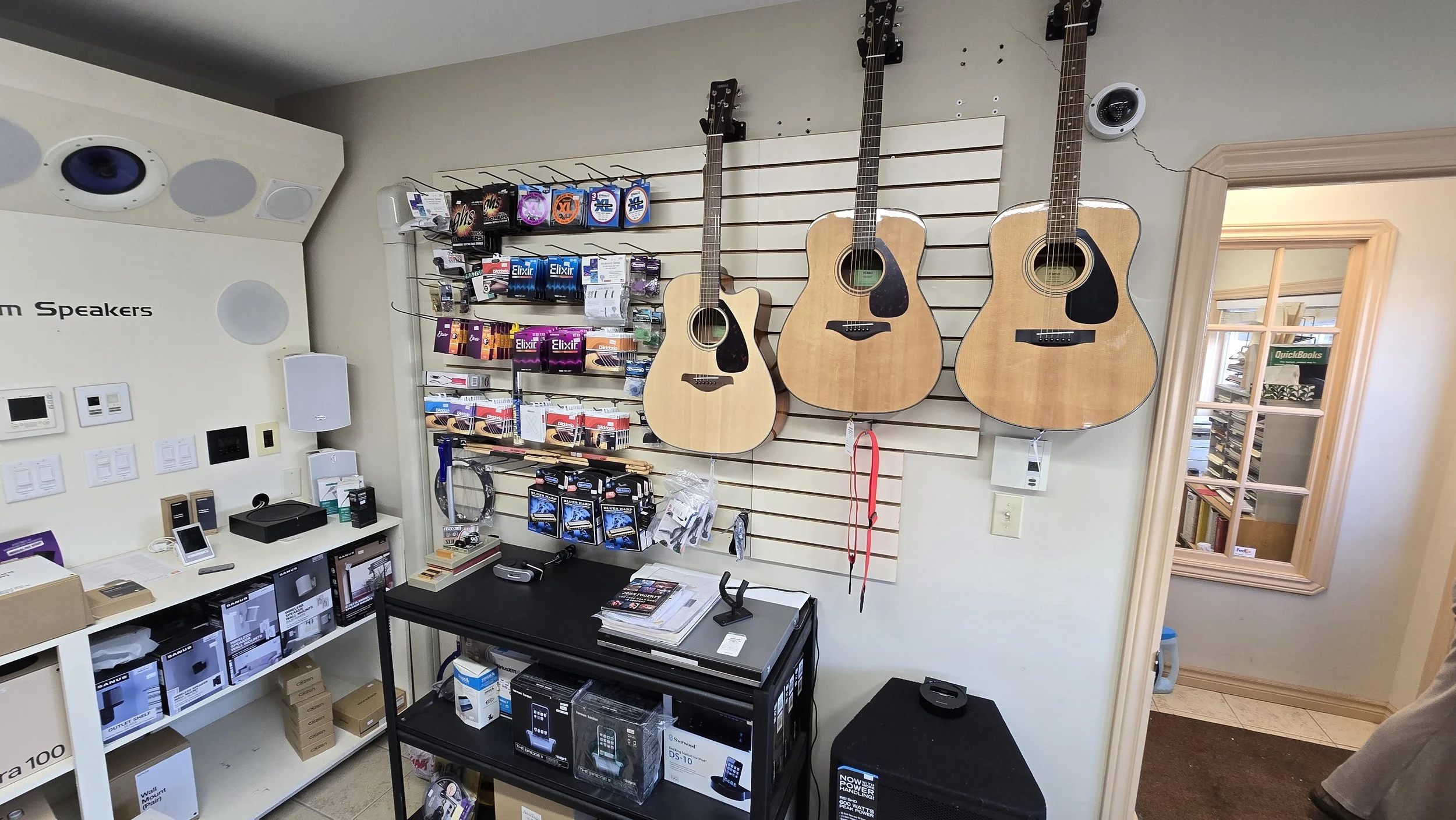 Three acoustic guitars hanging on a display wall in a music store. The store also has various guitar accessories, audio equipment, and boxes of electronics.