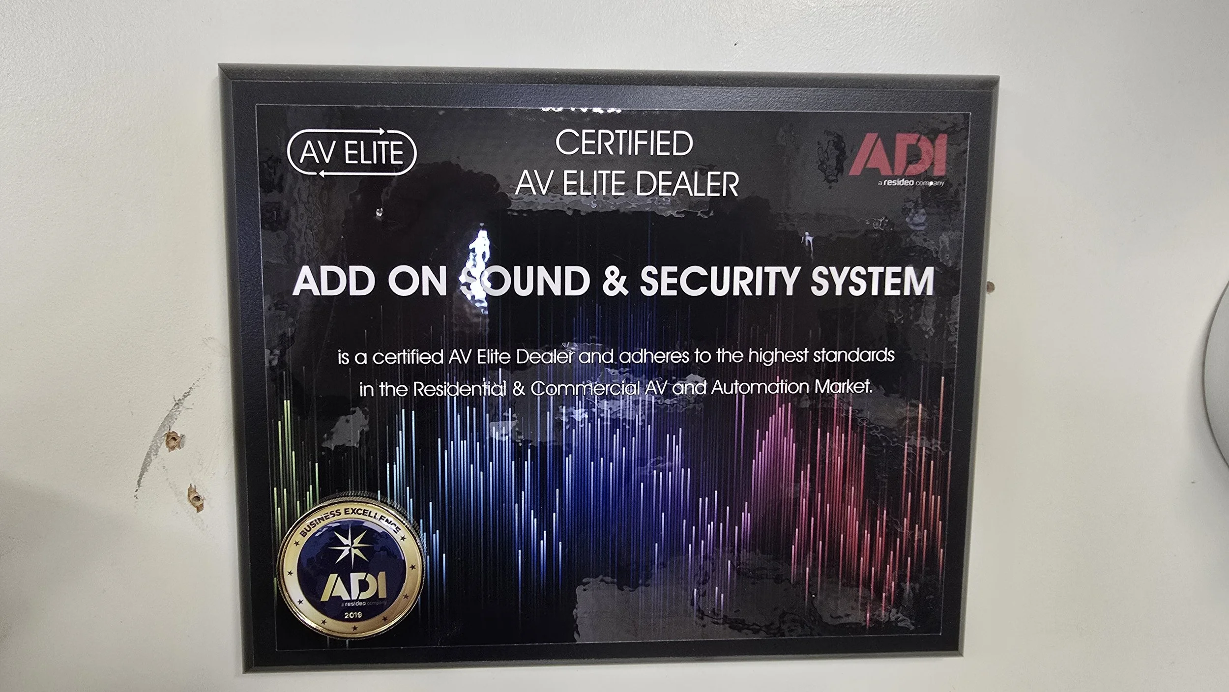 A certification plaque on a wall, indicating the business is a certified AV Elite Dealer specializing in sound and security systems, with a logo and colorful sound wave graphics.