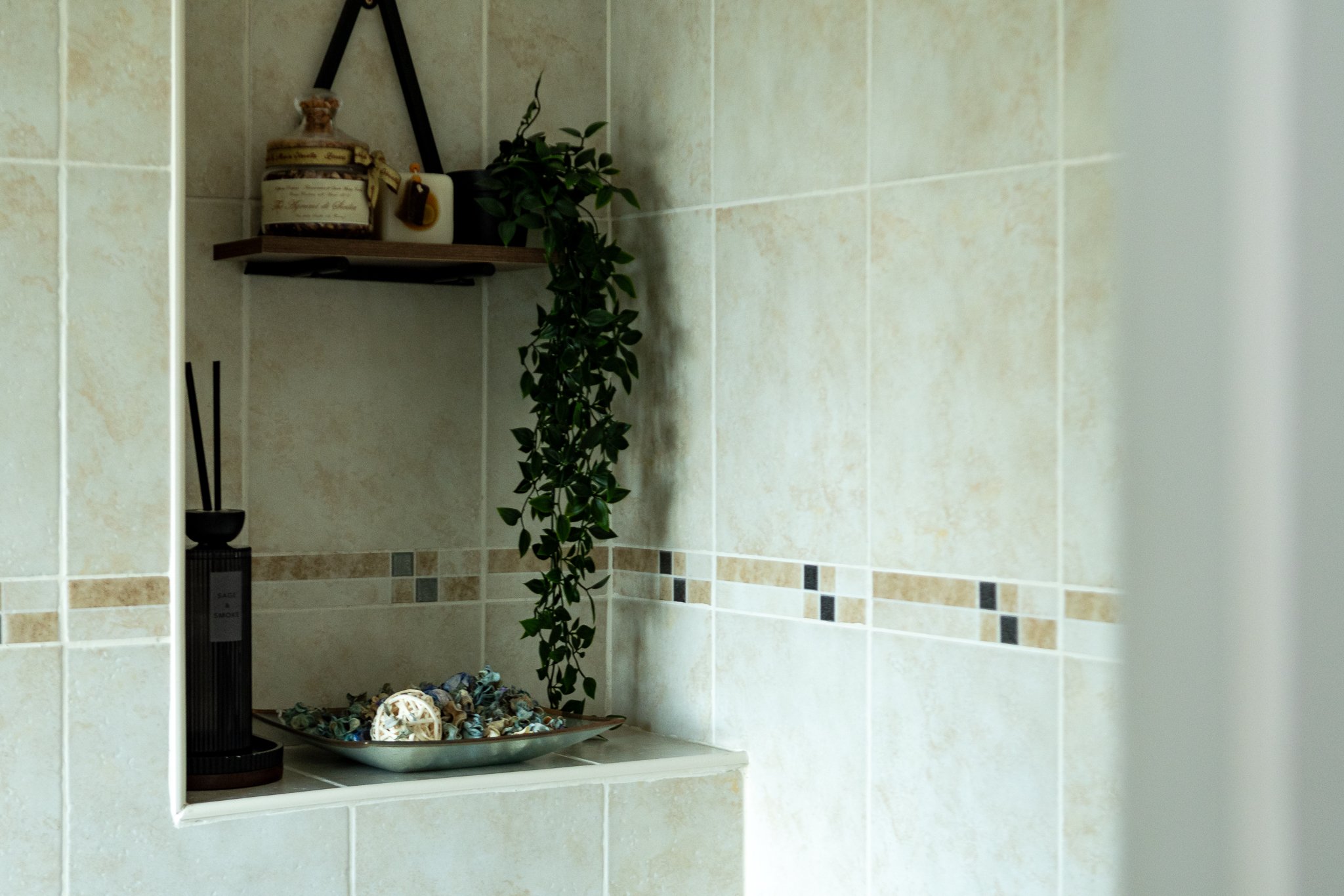 A bathroom nook with a shelf holding a decorative box and small container, a hanging green plant, and a tray of small stones and shells, all set against beige tiled walls.