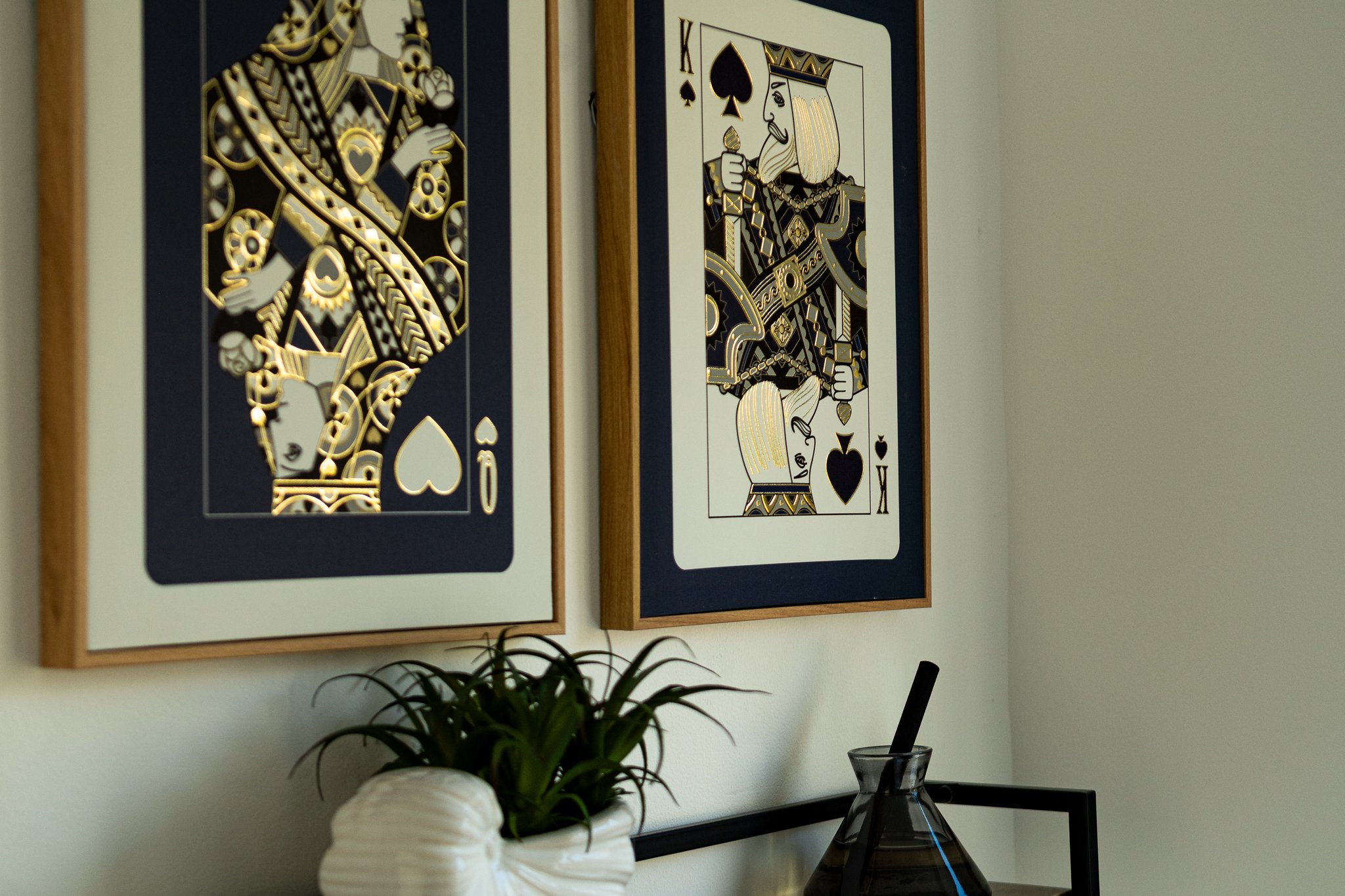 Decorative framed playing cards, the King of Spades and the Queen of Clubs, hanging on a wall above a plant and a glass container with a straw.