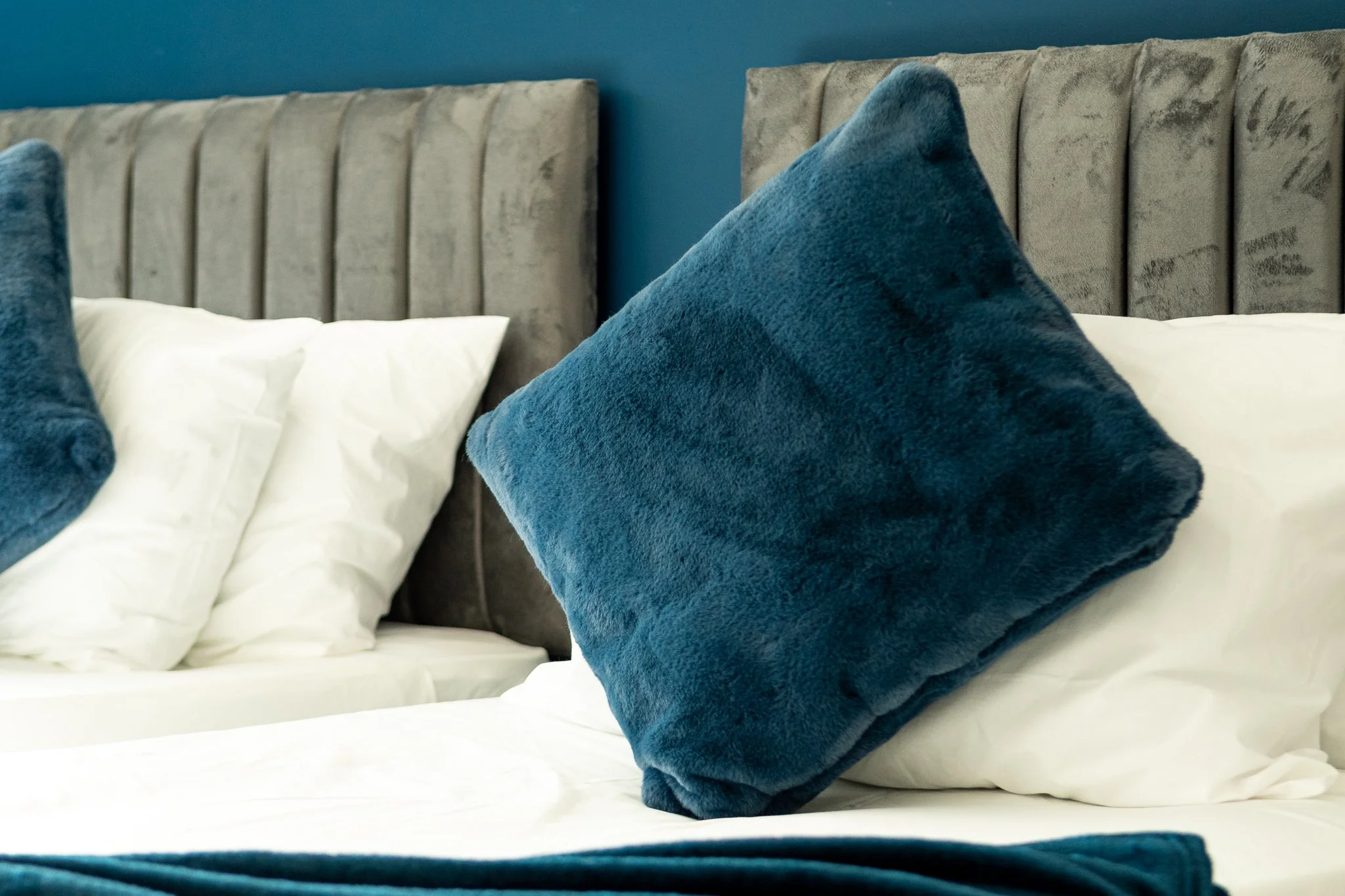 A close-up of a bed with white sheets, a gray upholstered headboard, and decorative blue pillows in front of a teal wall.