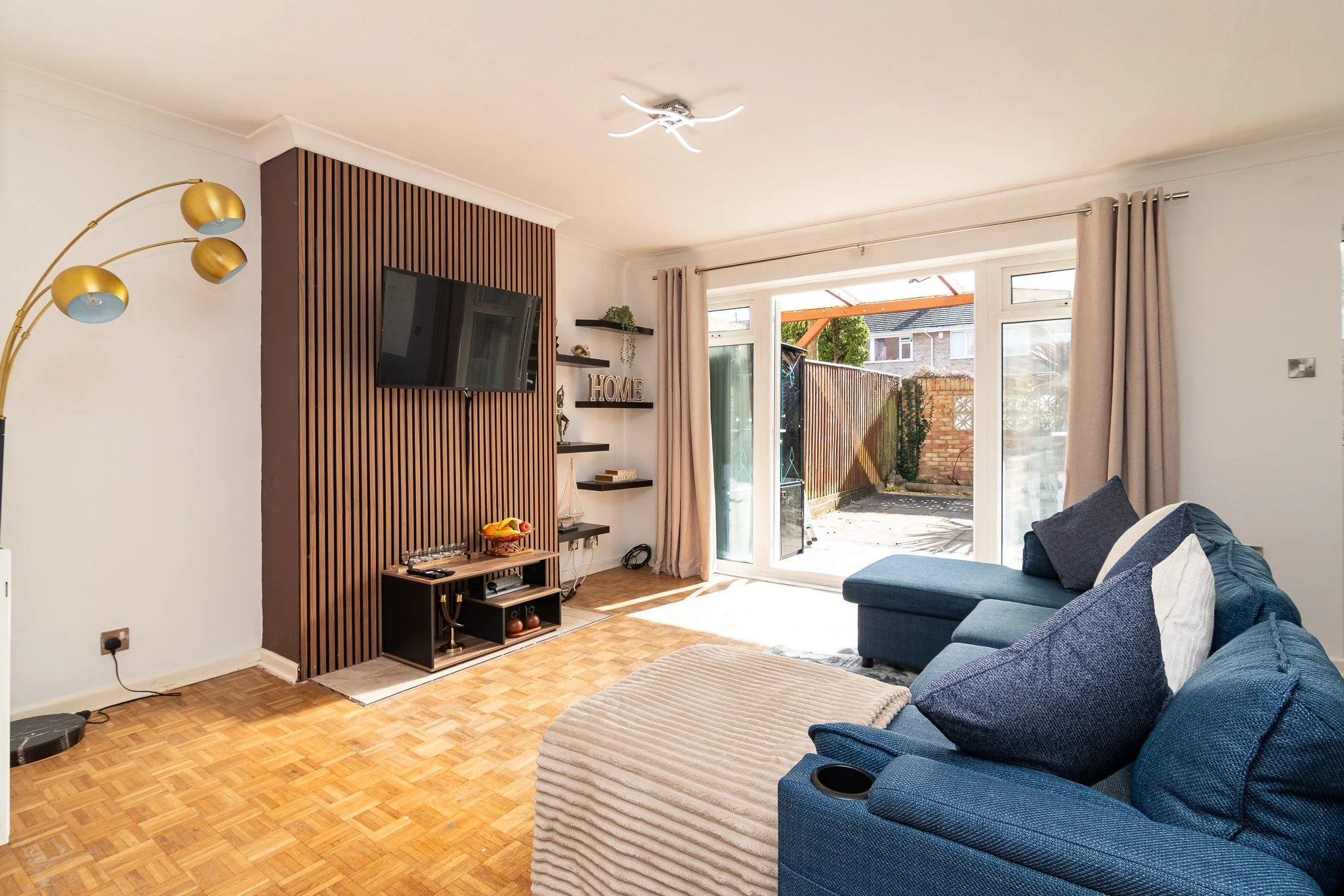 Living room with blue sectional sofa, beige ottoman, wooden flooring, TV on a wall with wooden panel, sliding glass doors to patio, beige curtains, floor lamp with gold accents, wall shelves with decor, backyard with brick wall and wooden fence