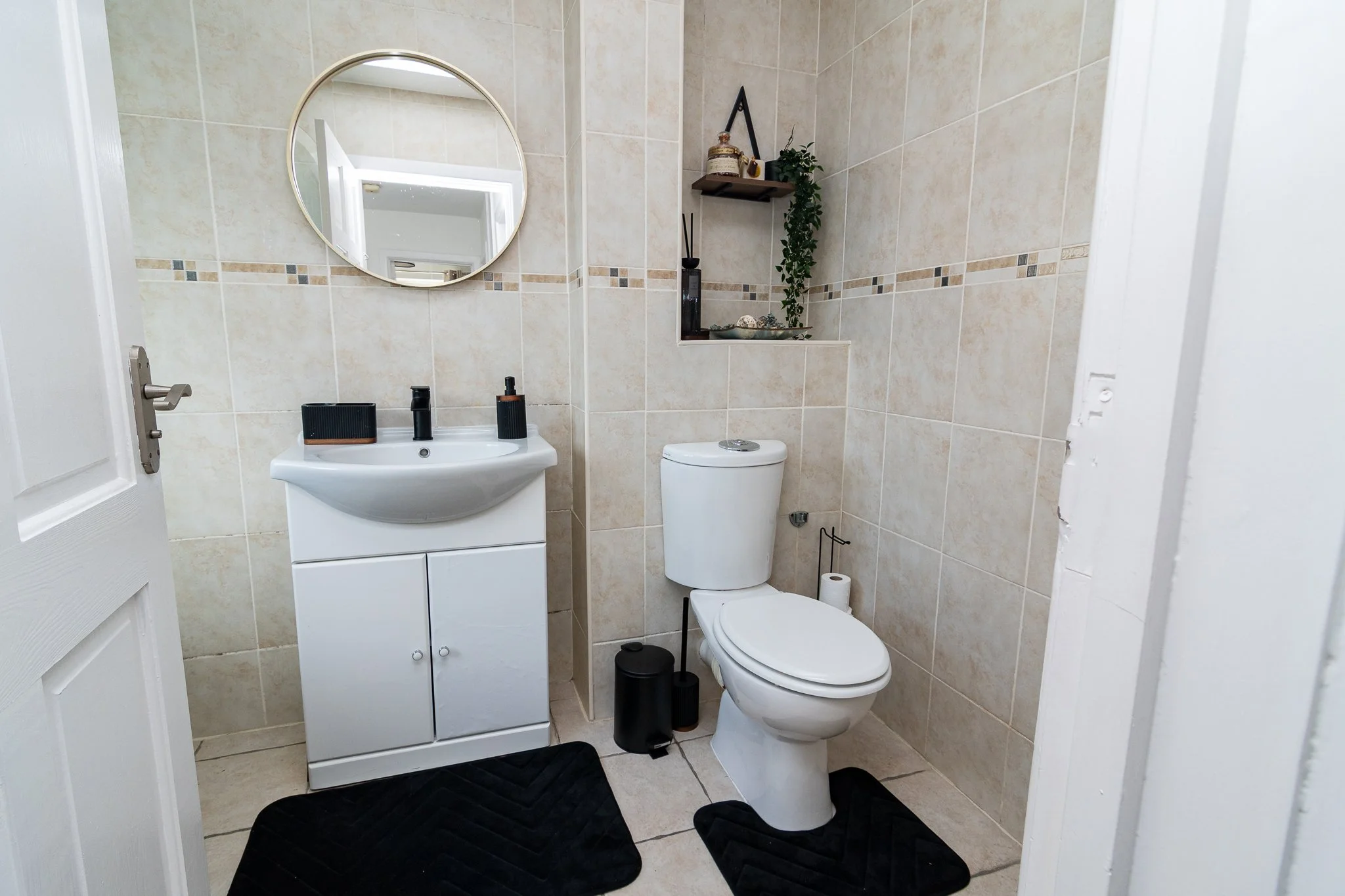 A small bathroom with beige tiled walls, a vanity with a mirror, and a toilet. Decor includes black accessories, small plants, and shelving with decorative items. Black rugs are on the floor.