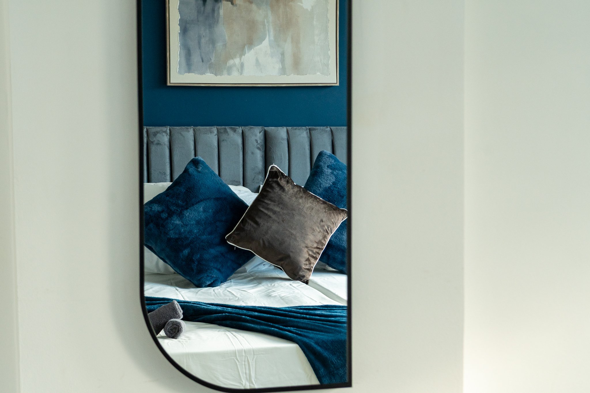 A mirror reflecting a neatly made bed with white sheets, a velvet headboard, and decorative pillows in dark blue and brown. There is a folded towel on the corner of the bed and a blue throw blanket.