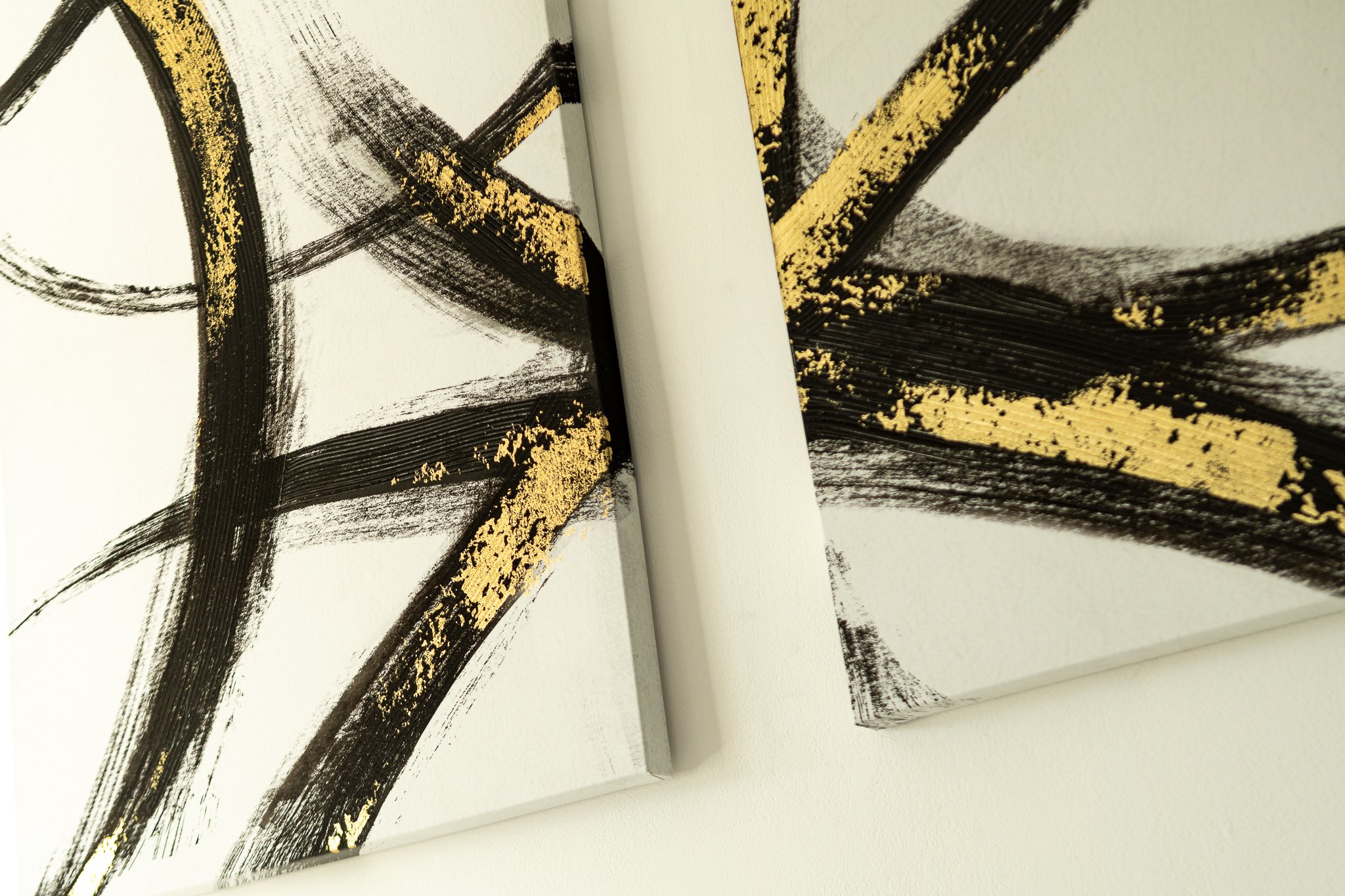 An abstract painting featuring bold black brushstrokes with gold accents on a white canvas, positioned overlapping each other.