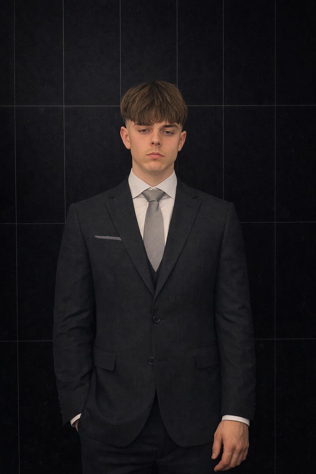 A young man in a dark suit, white shirt, and light gray tie standing against a black tiled wall.