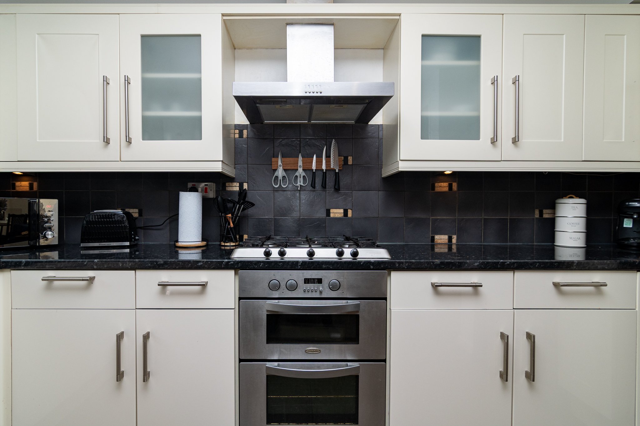 Modern kitchen with white cabinets, black countertops, stainless steel oven, and black tiled backsplash with decorative gold accents. Items include knives, scissors, paper towel, toaster, and spice containers.