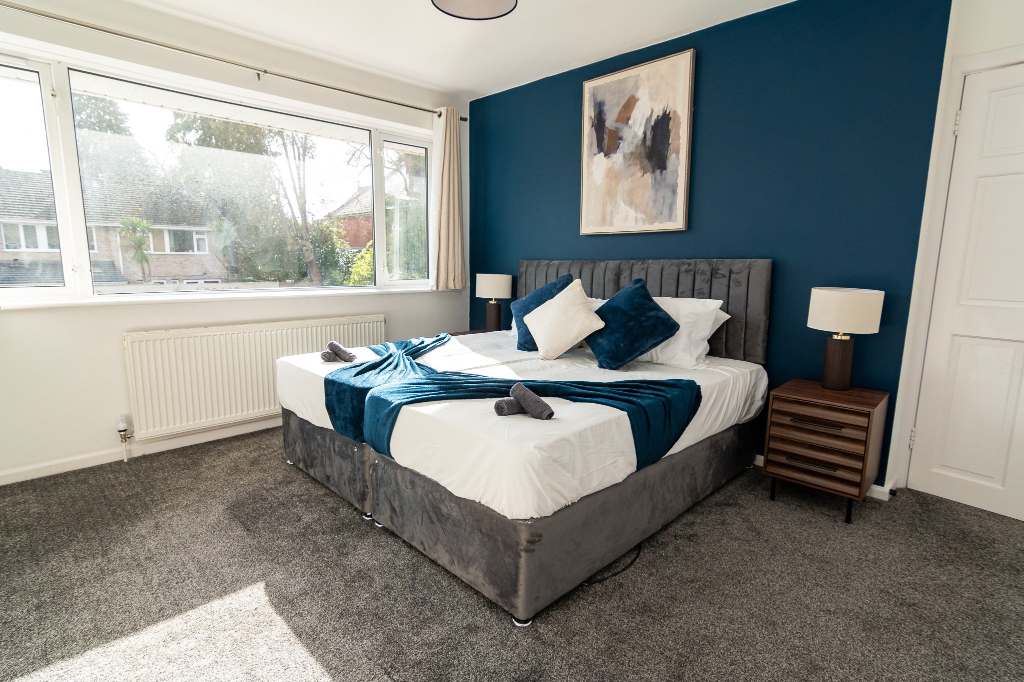 A bedroom with a large window, a bed with a blue and white color scheme, a gray velvet headboard, and wooden nightstands with lamps.