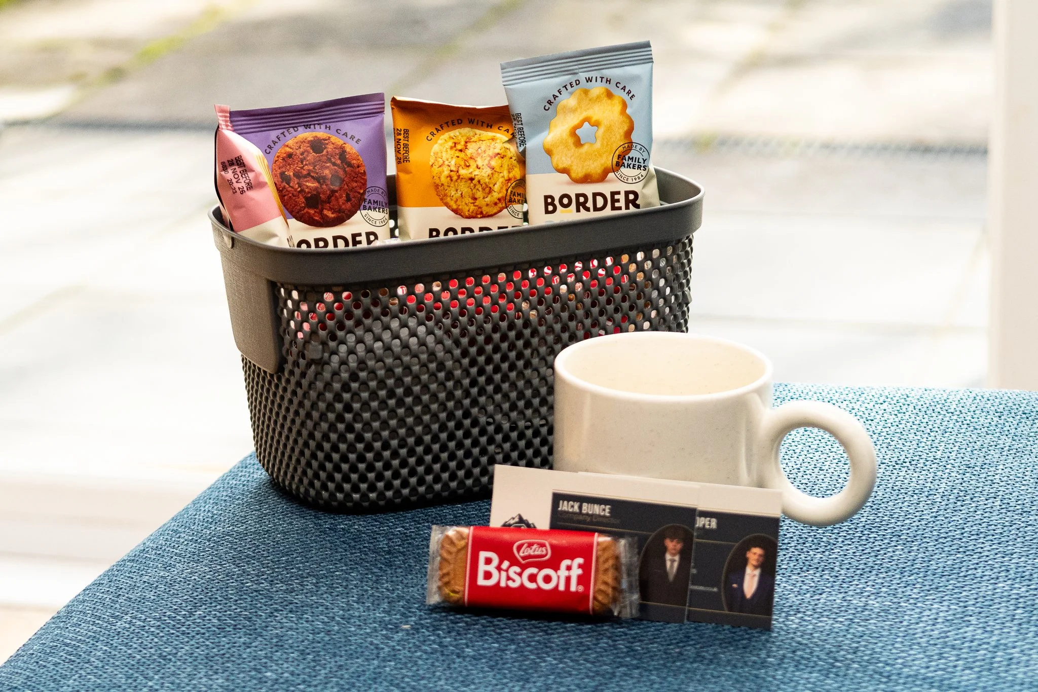 A black perforated basket containing three snack bags of cookies with colorful packaging, placed on a blue fabric surface next to a white coffee mug, a Lotus Biscoff cookie in a red wrapper, and a card featuring a person in a tuxedo, all set against a bright background with a blurry outdoor scene.