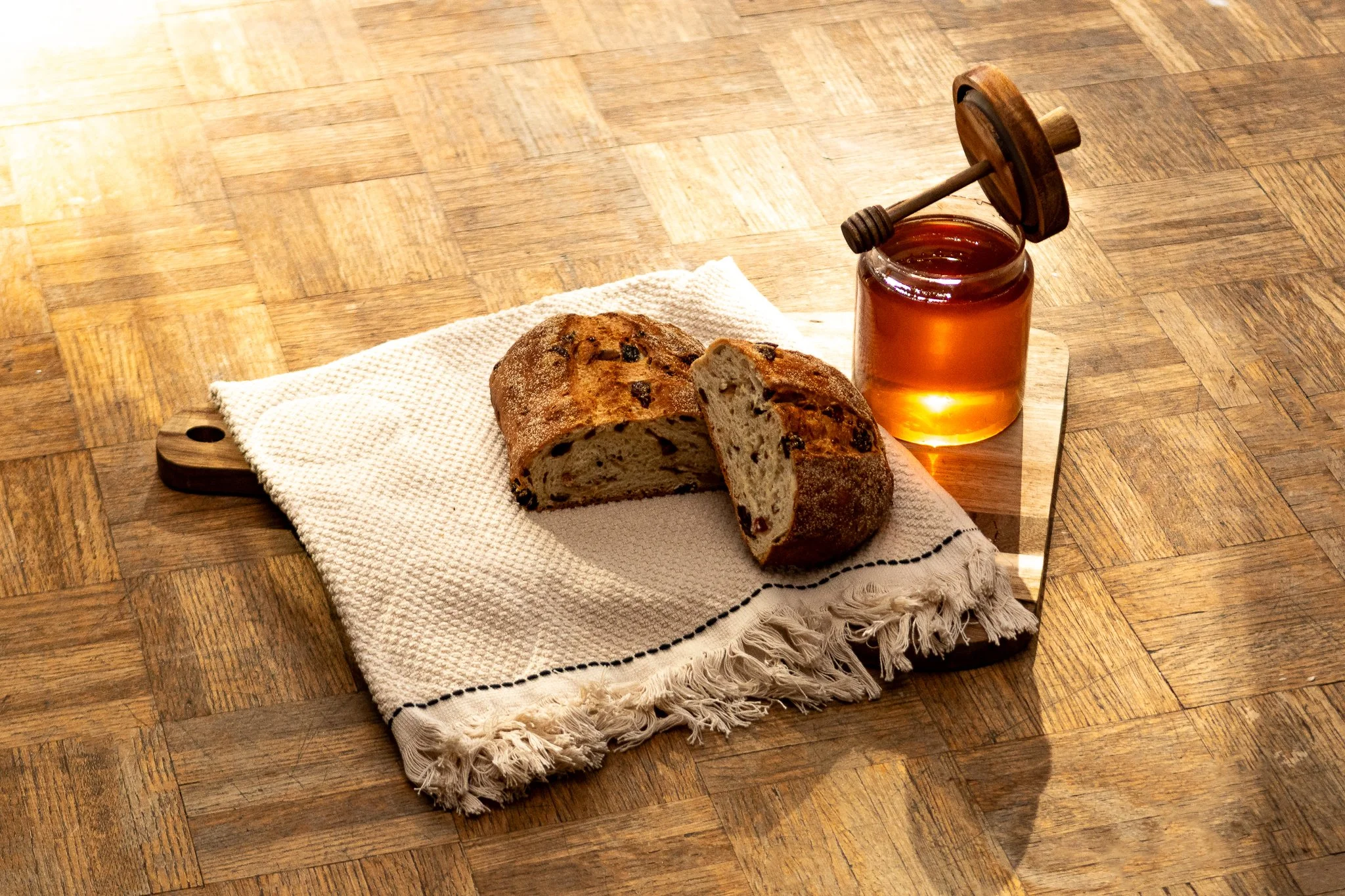 A loaf of raisin bread with a slice cut and placed on a white cloth on a wooden cutting board, next to a jar of honey with a honey dipper resting on top, on a wooden table.