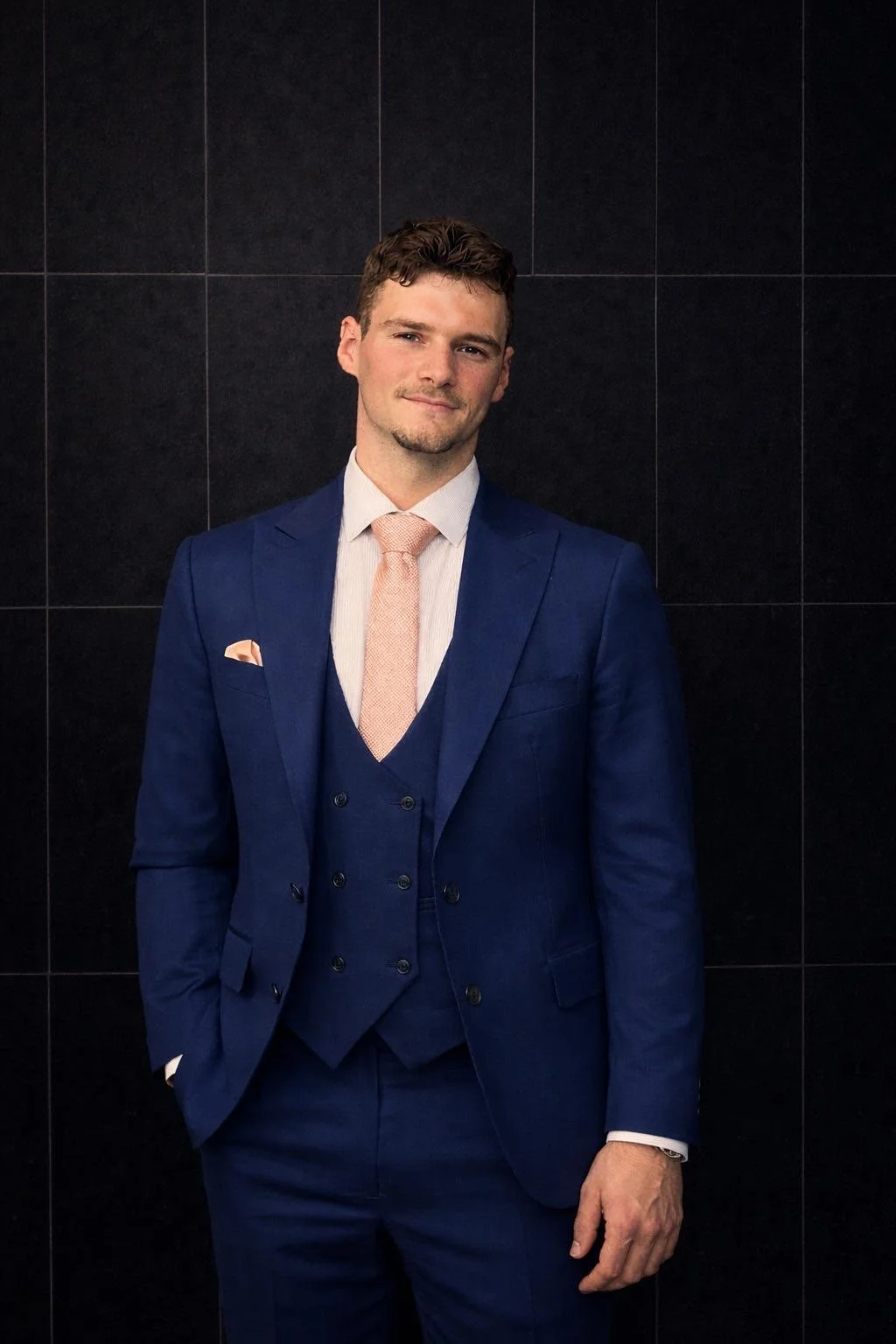 A young man in a blue suit standing against a black tiled wall, smiling slightly, with his right hand in his pocket.