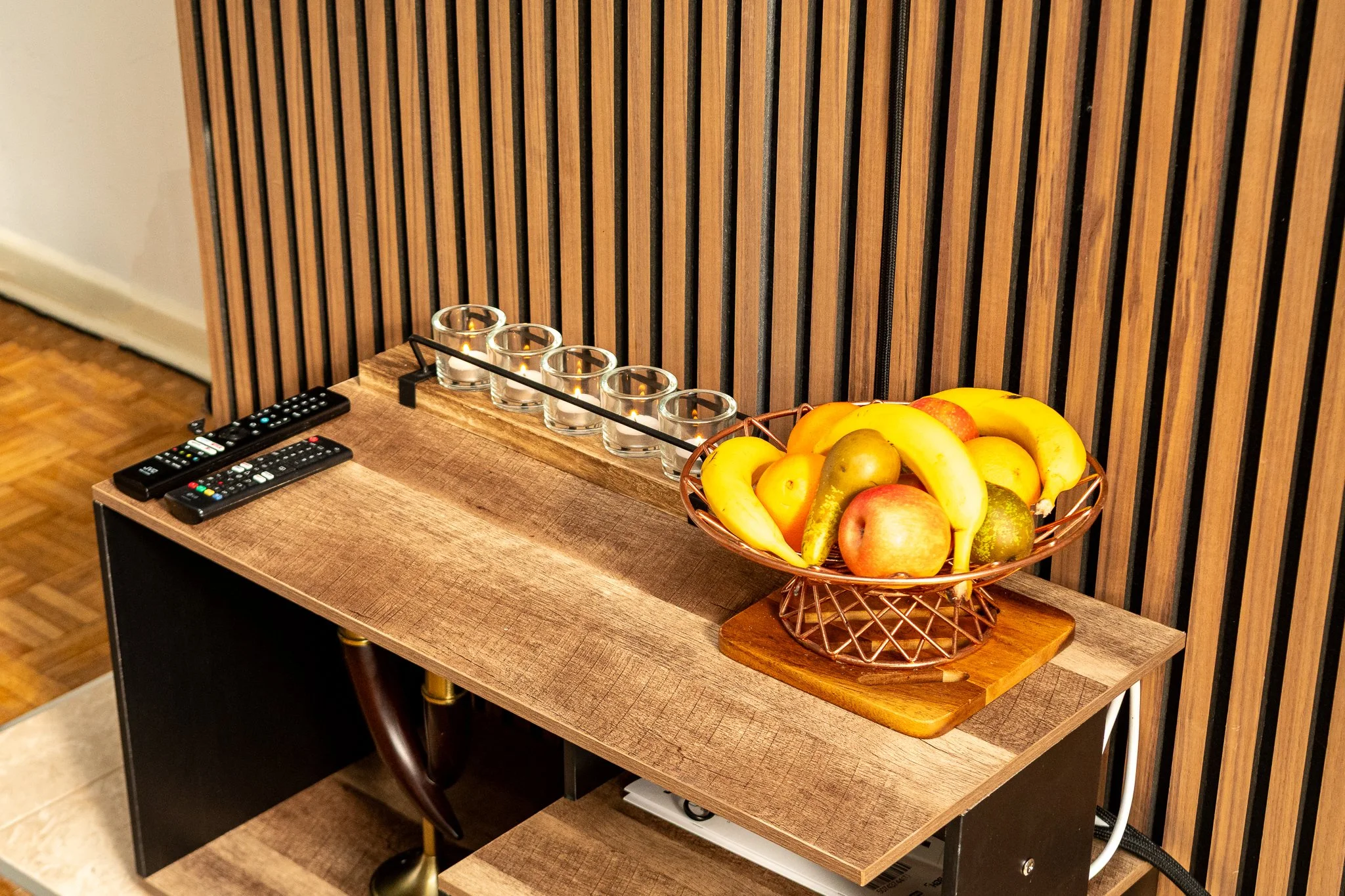 A wooden side table with a bowl of bananas, apples, and pears on a small cutting board, four empty glass candles, and two remote controls, with a wooden slat wall in the background.