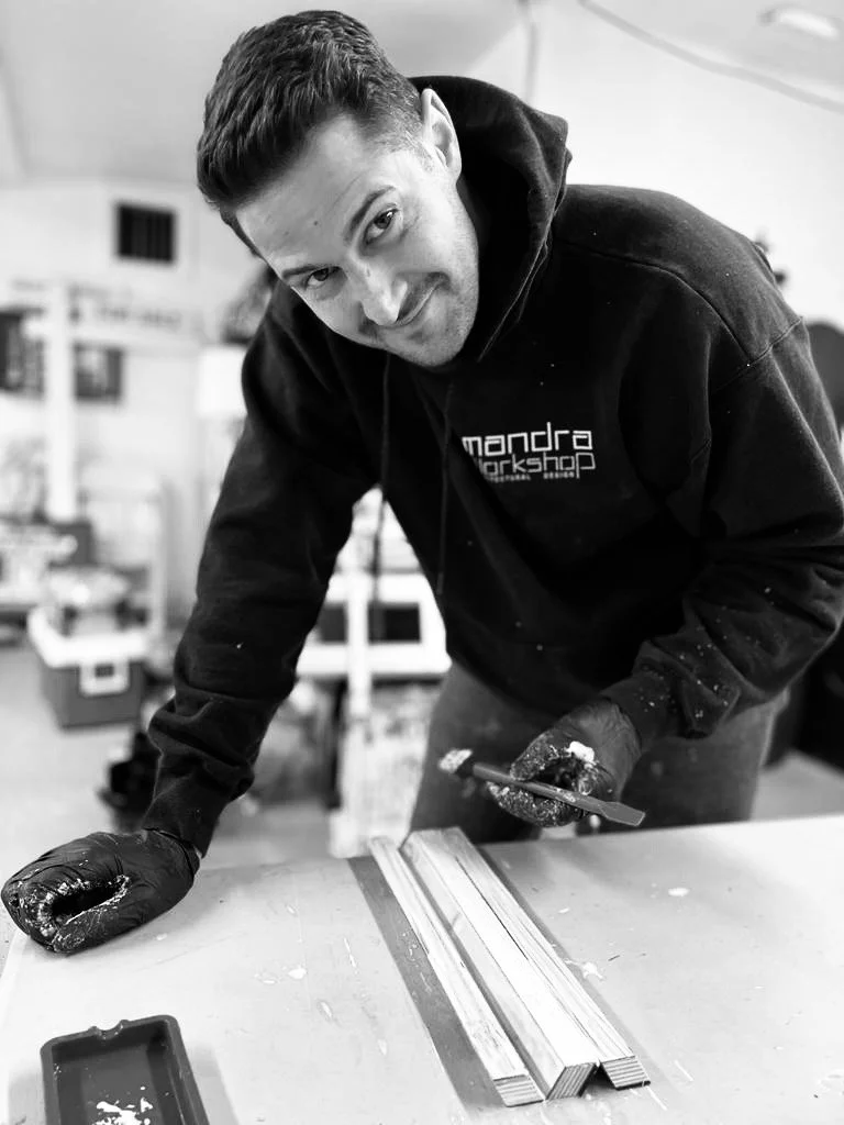 A man wearing a hoodie and gloves leans over a workbench with a smile, working on a woodworking project in a workshop.