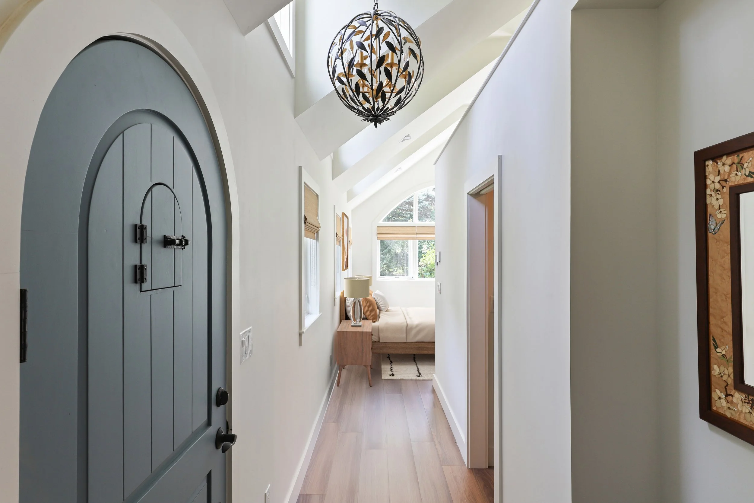 A bright hallway leading to a bedroom with a large window, white walls, wooden flooring, and a decorative ceiling light fixture.