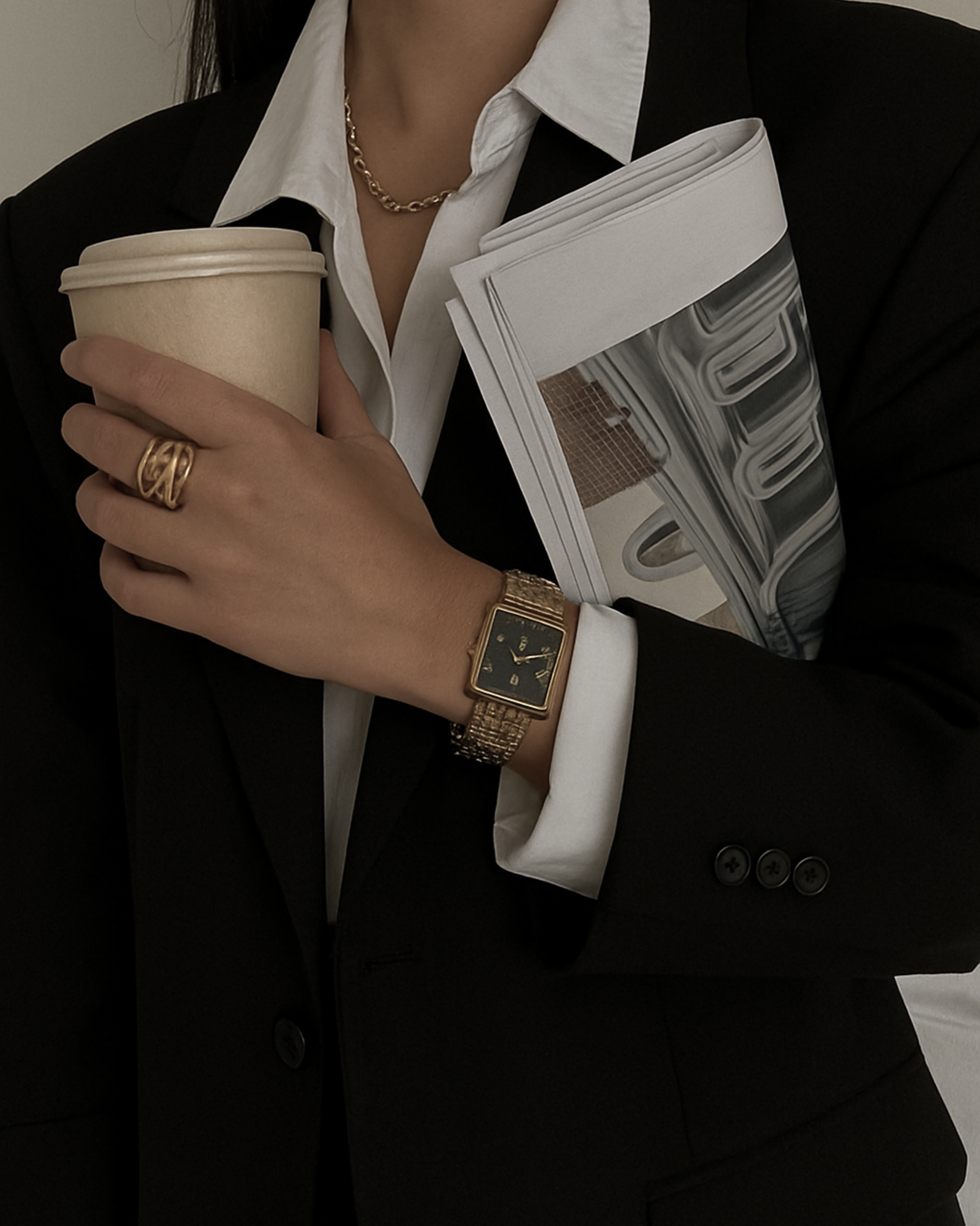 A woman in a black blazer and white shirt holding a paper coffee cup, a stack of documents, and wearing a gold watch, ring, and necklace.