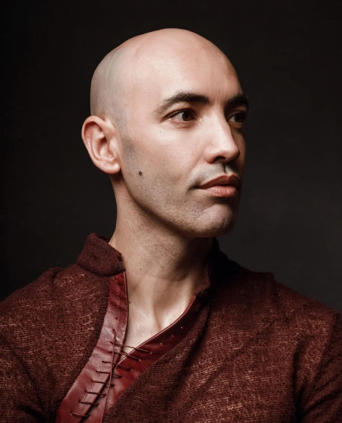 Portrait of a bald man wearing a maroon textured jacket, looking to the right, against a dark background.