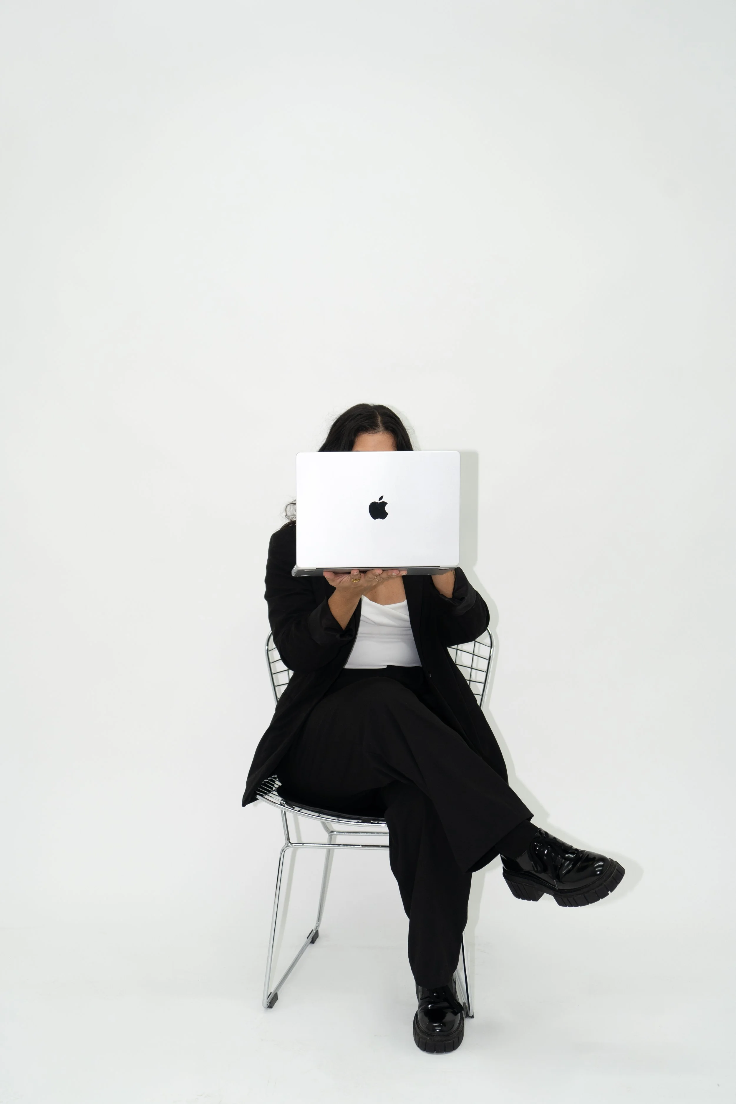 Person sitting on a wire chair using a silver MacBook in a plain white room.