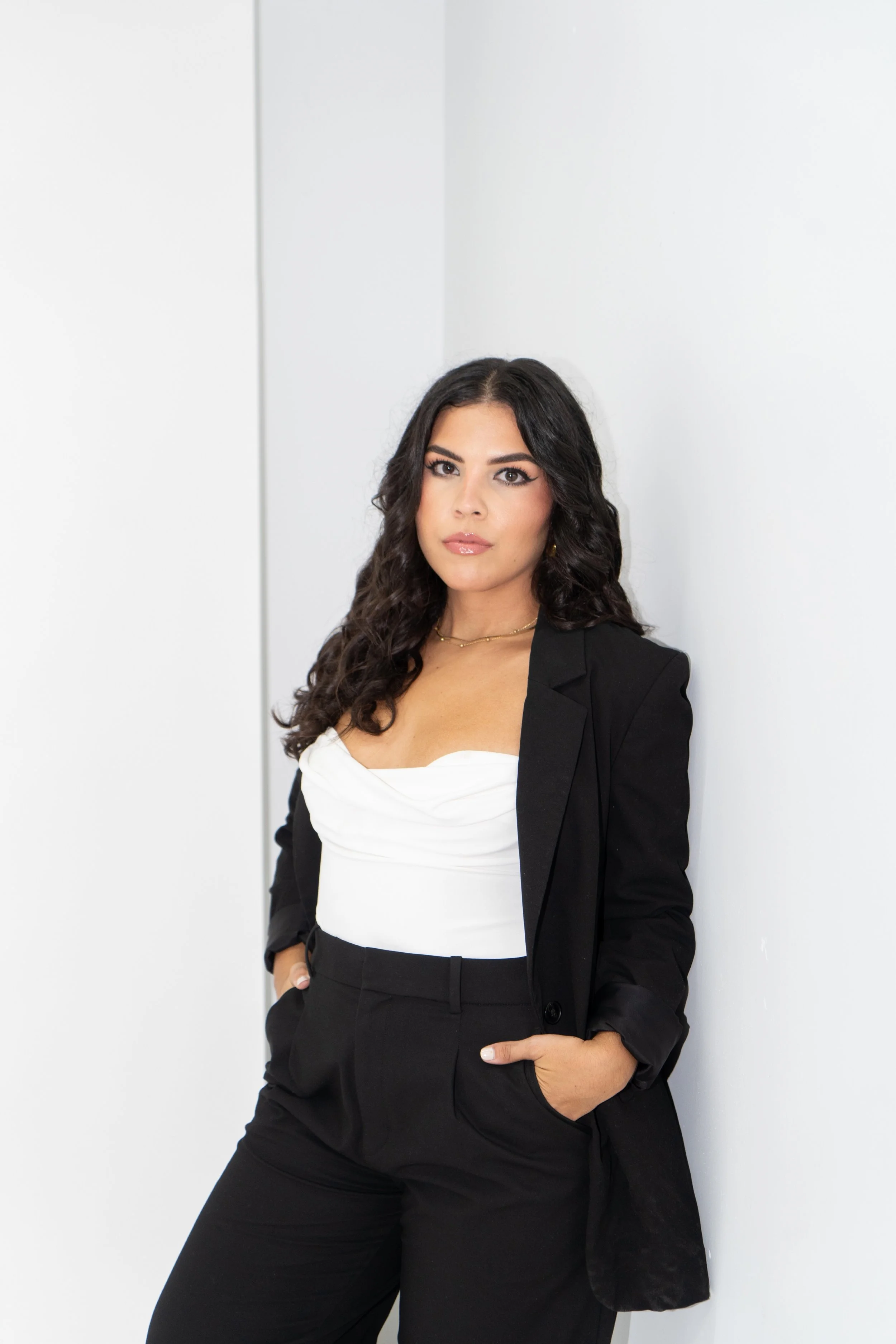 A woman with dark, curly hair wearing a black blazer and pants, white top, posing against a white wall.