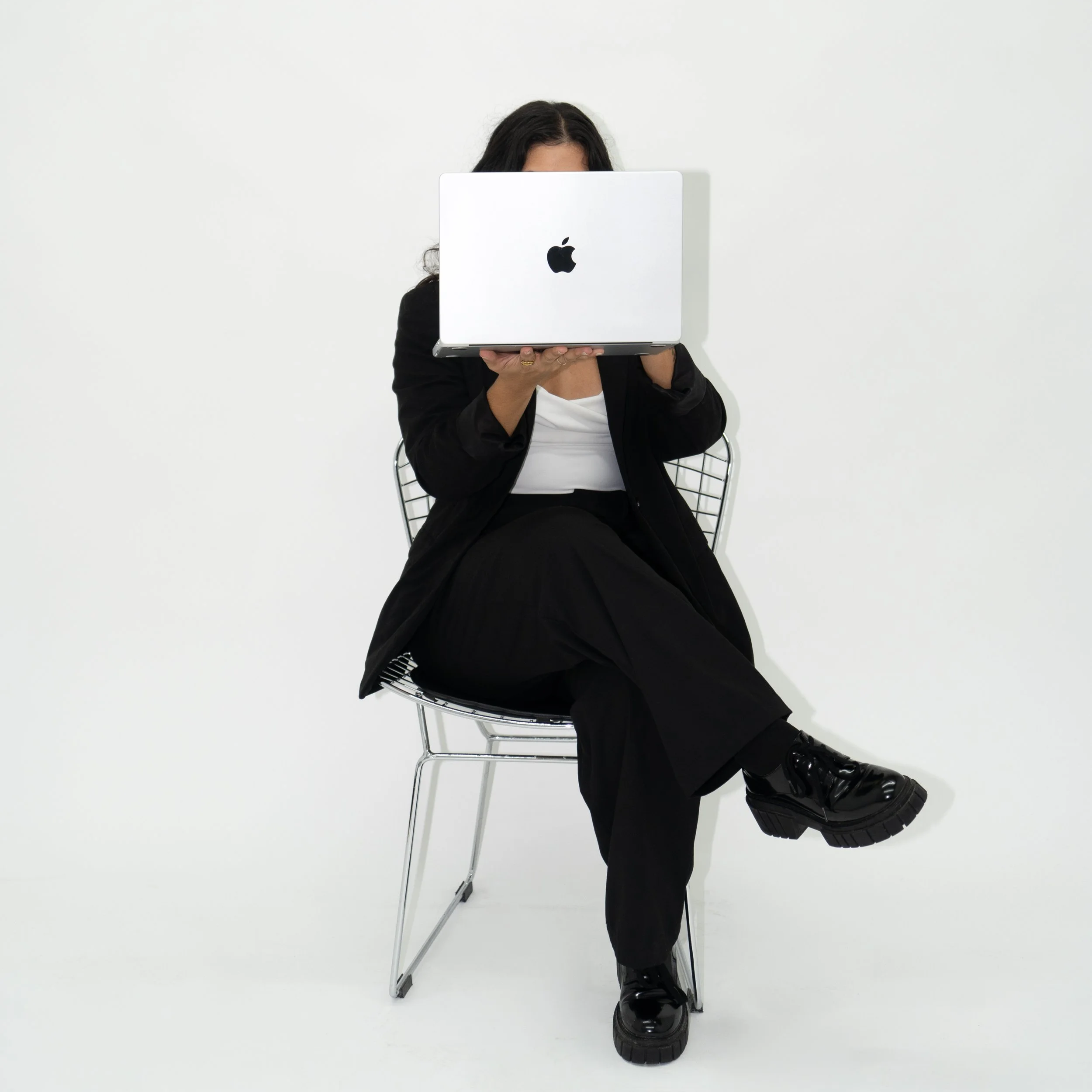 Person sitting on a modern wire chair, holding a silver Apple MacBook in front of face, against a plain white background.