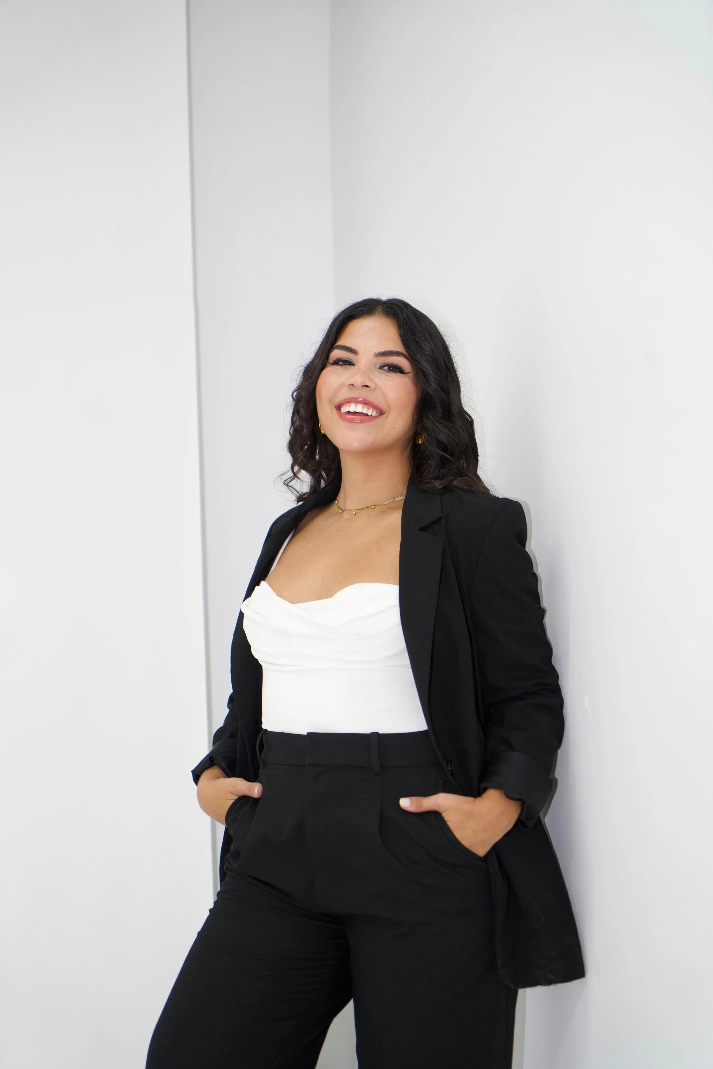 A woman in a black blazer and pants with a white top, standing against a plain white wall, smiling and looking at the camera.