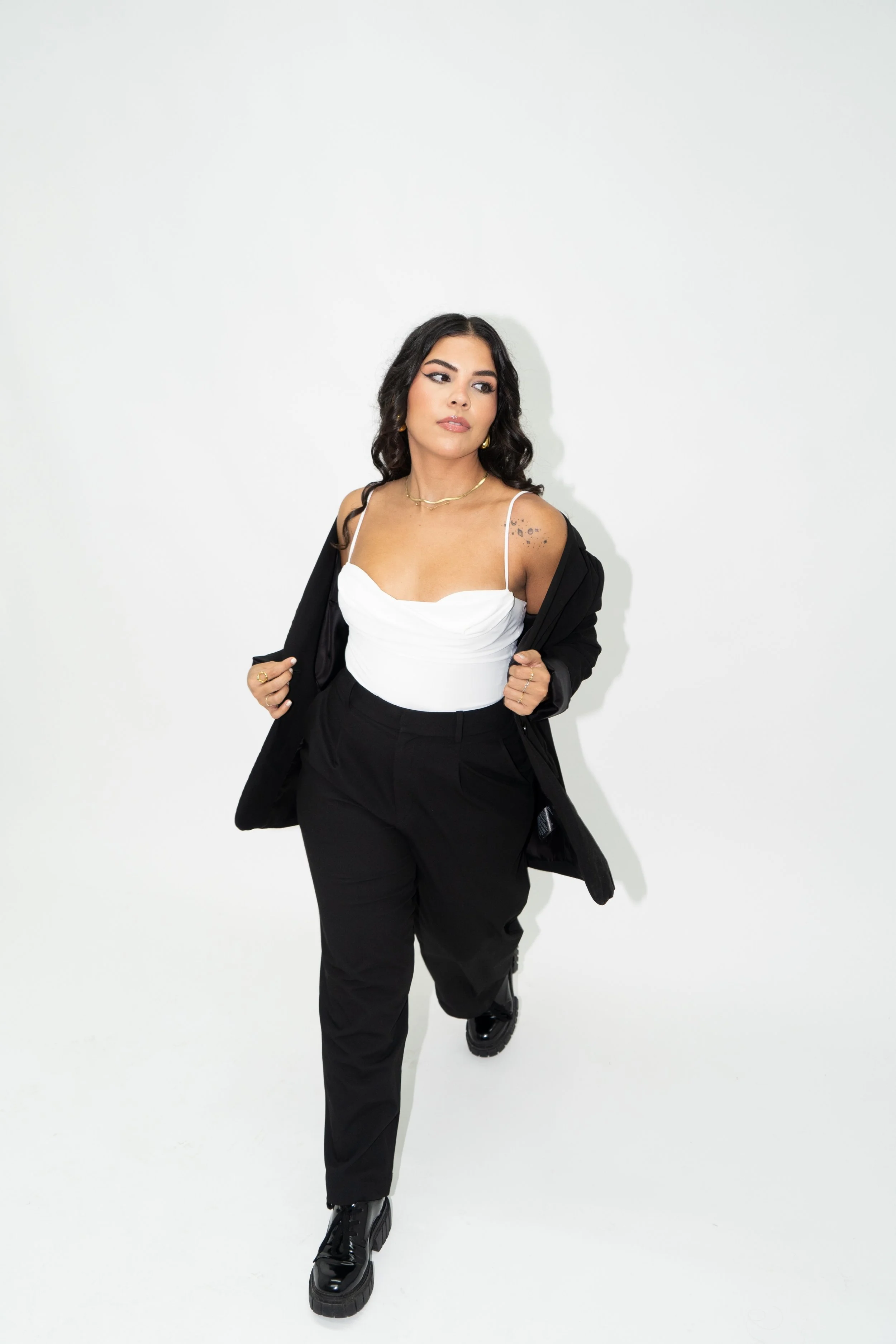 A woman with dark wavy hair, dressed in black pants, a white top, and a black jacket, is walking towards the camera against a plain white background.
