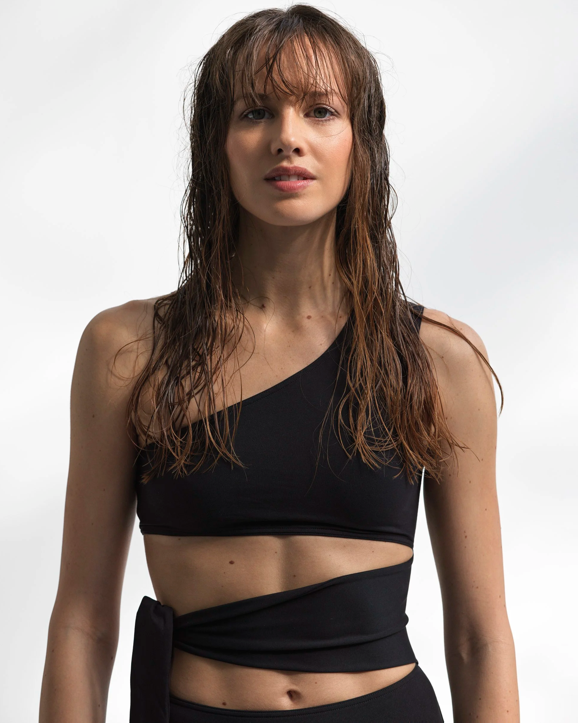 A woman with long, wet, brown hair, wearing a black one-shoulder crop top and a matching wrap skirt, standing against a plain white background.