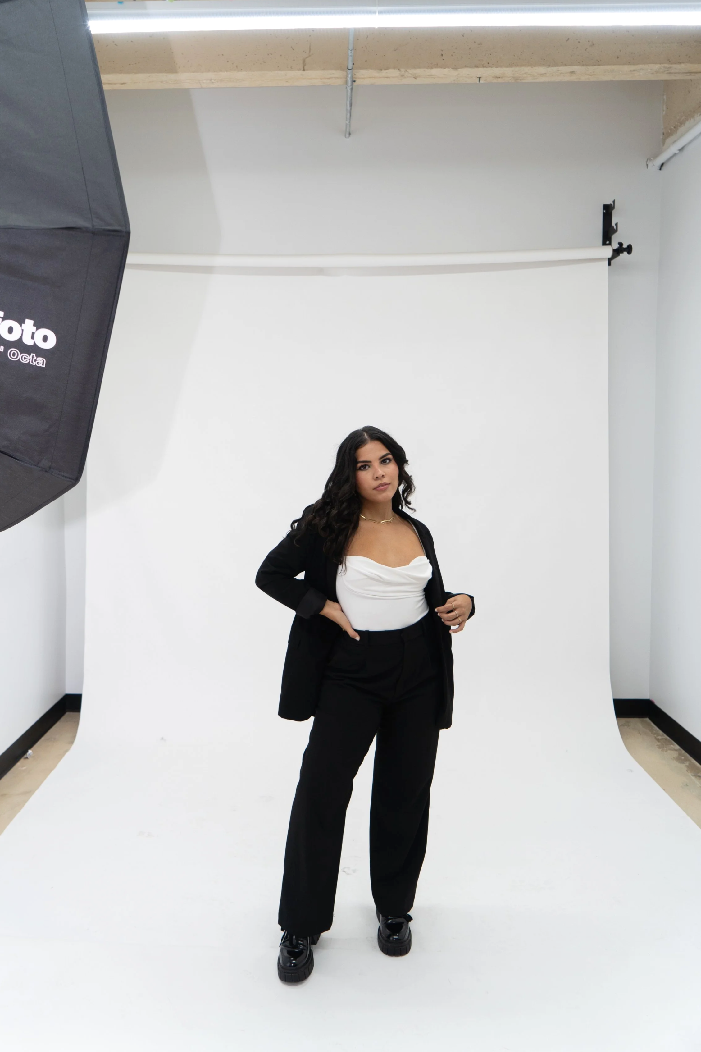 A woman standing in a photography studio backdrop, wearing a black blazer, white top, matching black pants, and black shoes, with professional lighting equipment visible.