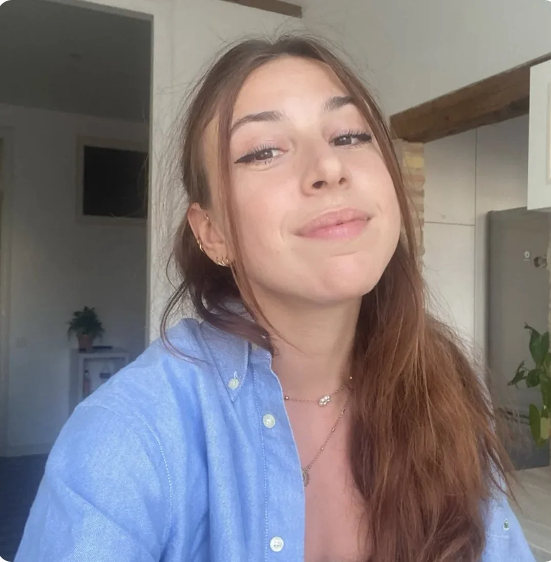 Une jeune femme avec des cheveux bruns et longs, portant une chemise en jean bleu, souriant légèrement, en intérieur avec mur blanc, télévision et plantes en arrière-plan.