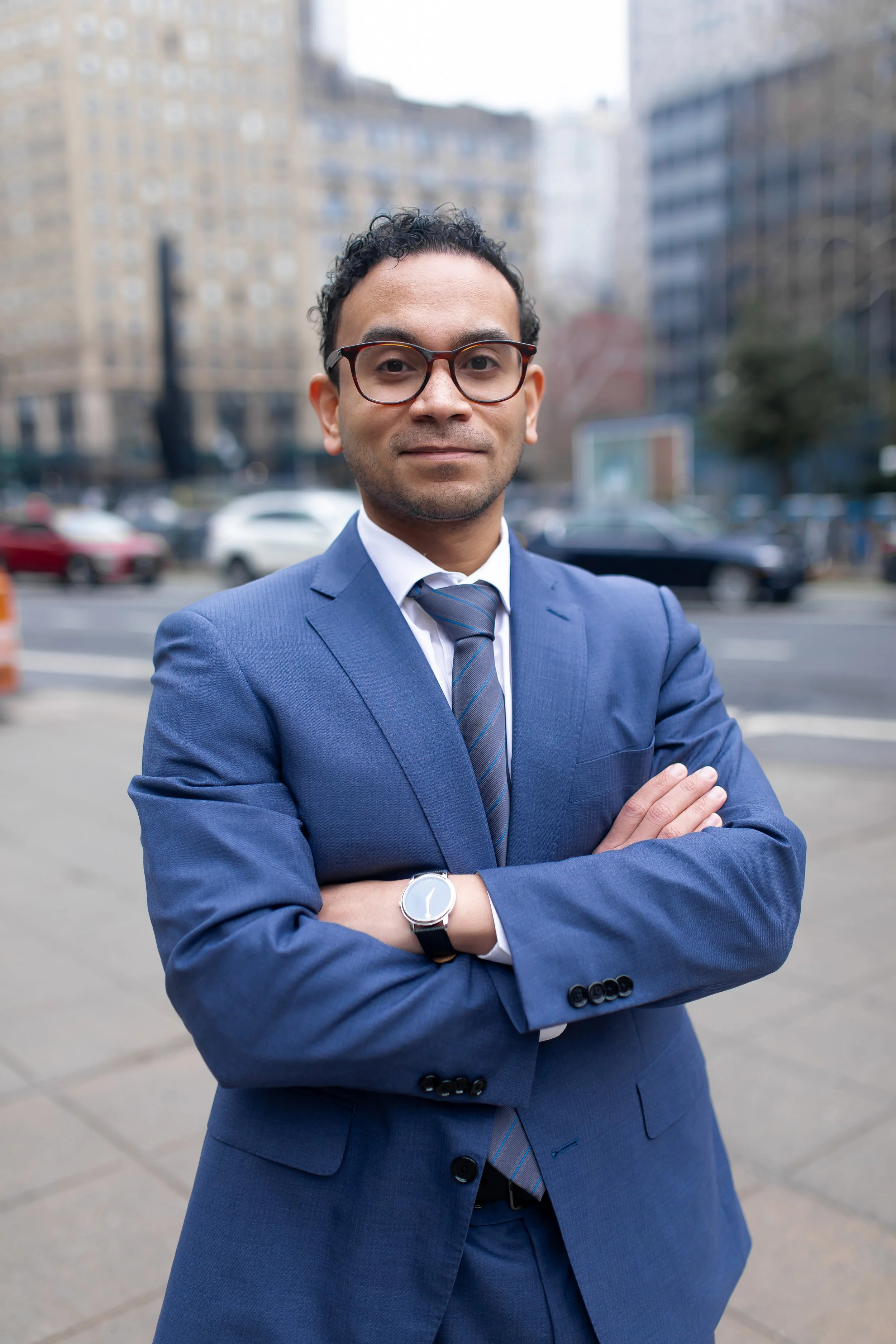 man in a blue suit with dark glasses standing with arms folded across chest