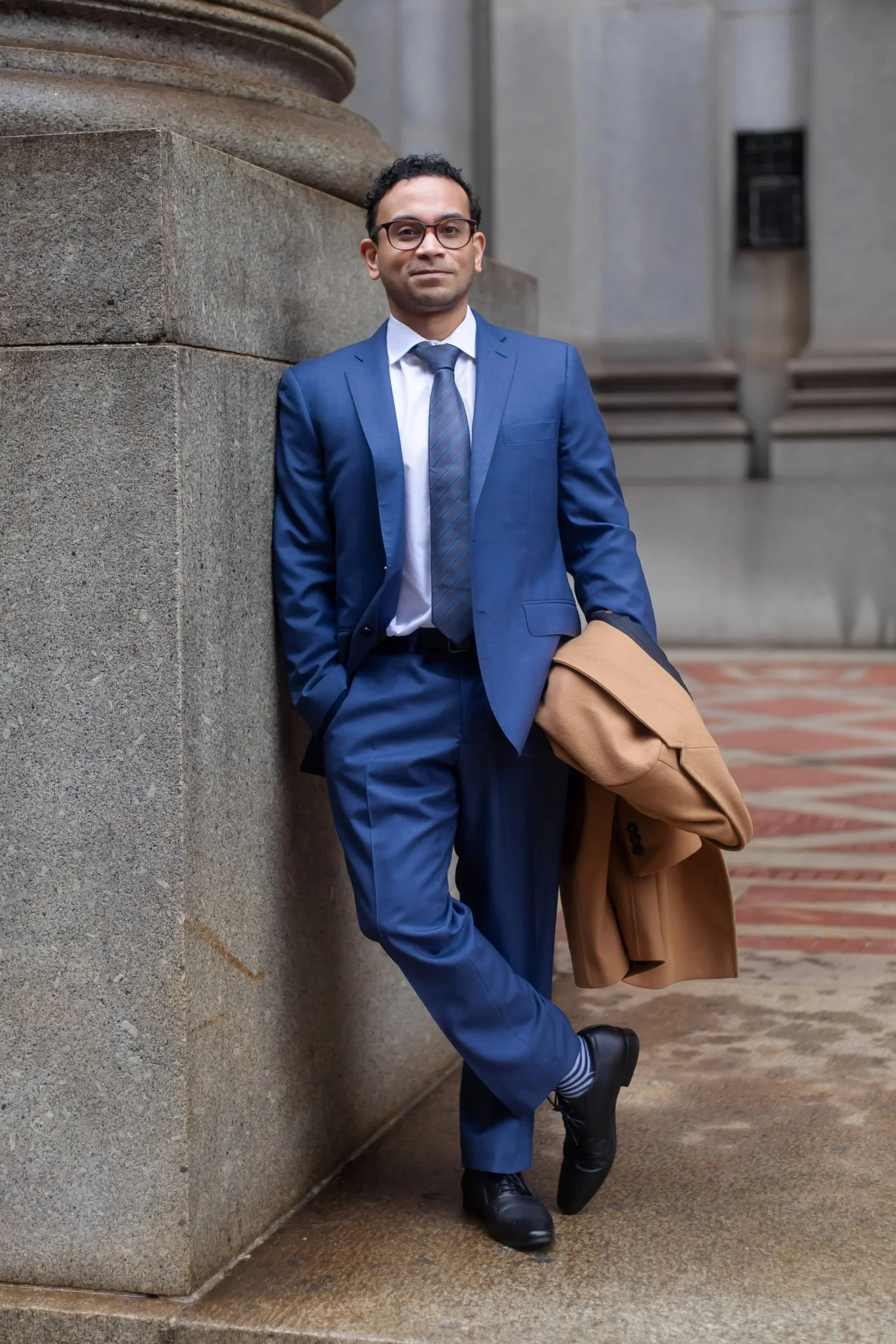 man in a blue suit with dark glasses holding a camel coat, standing with his leg crossed at his ankle and hand in his pocket