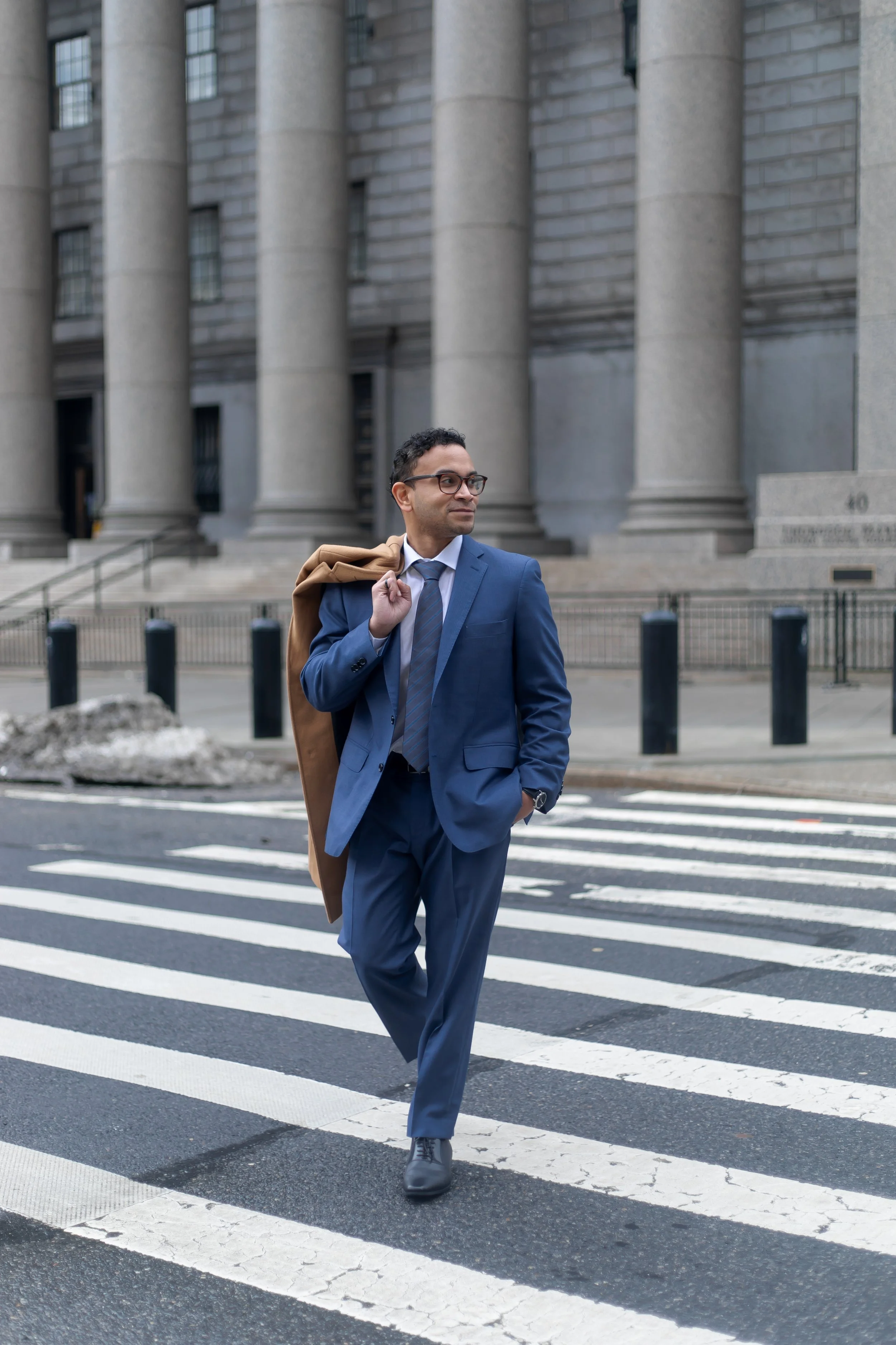 man in a blue suit with glasses and camel coat over his shoulder walking over a zebra crossing
