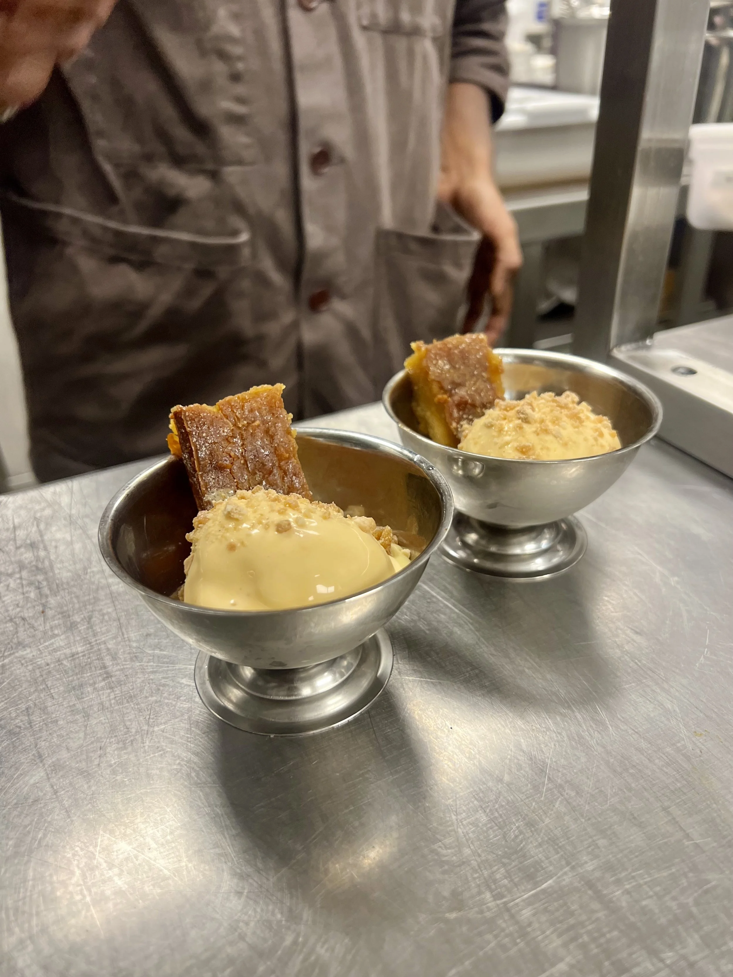 Two metal cups of vanilla ice cream with caramel sauce and a piece of cake on top, placed on a stainless steel countertop in a kitchen setting.