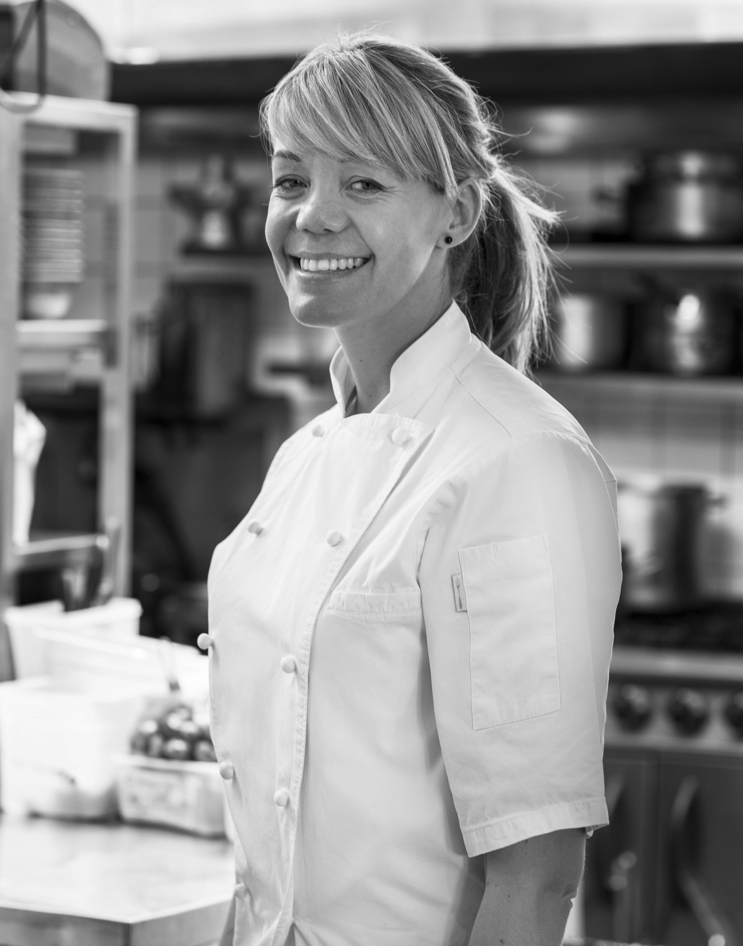 A smiling female chef in a professional kitchen, wearing a white chef's coat with shelves of cookware behind her.