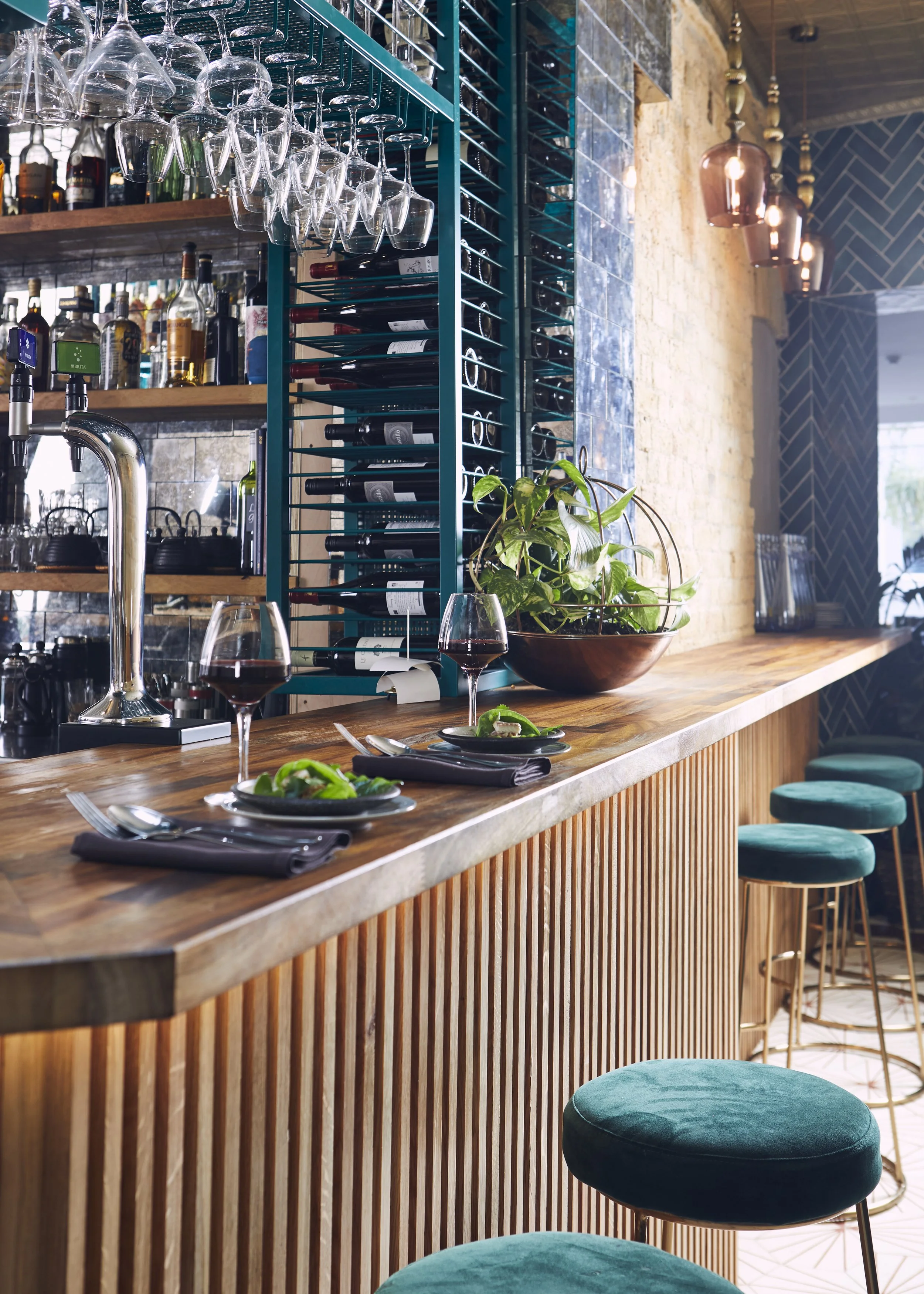 A cozy bar area with a wooden counter set for dining, featuring two plates with salads, two glasses of red wine, and set utensils. The bar's background shows hanging glasses, shelves with bottled spirits, and a wine rack with bottles. Decor includes a large plant in a bowl and modern hanging pendant lights, with teal upholstered bar stools lined up along the counter.