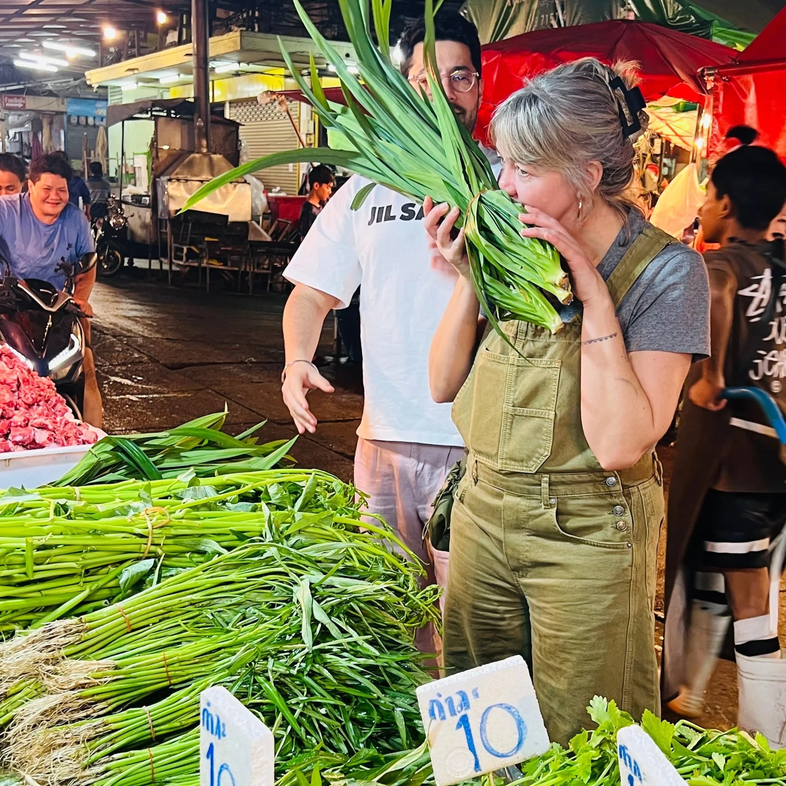 A woman smelling a bunch of green leafy vegetables at an outdoor market, with other shoppers and market stalls in the background.